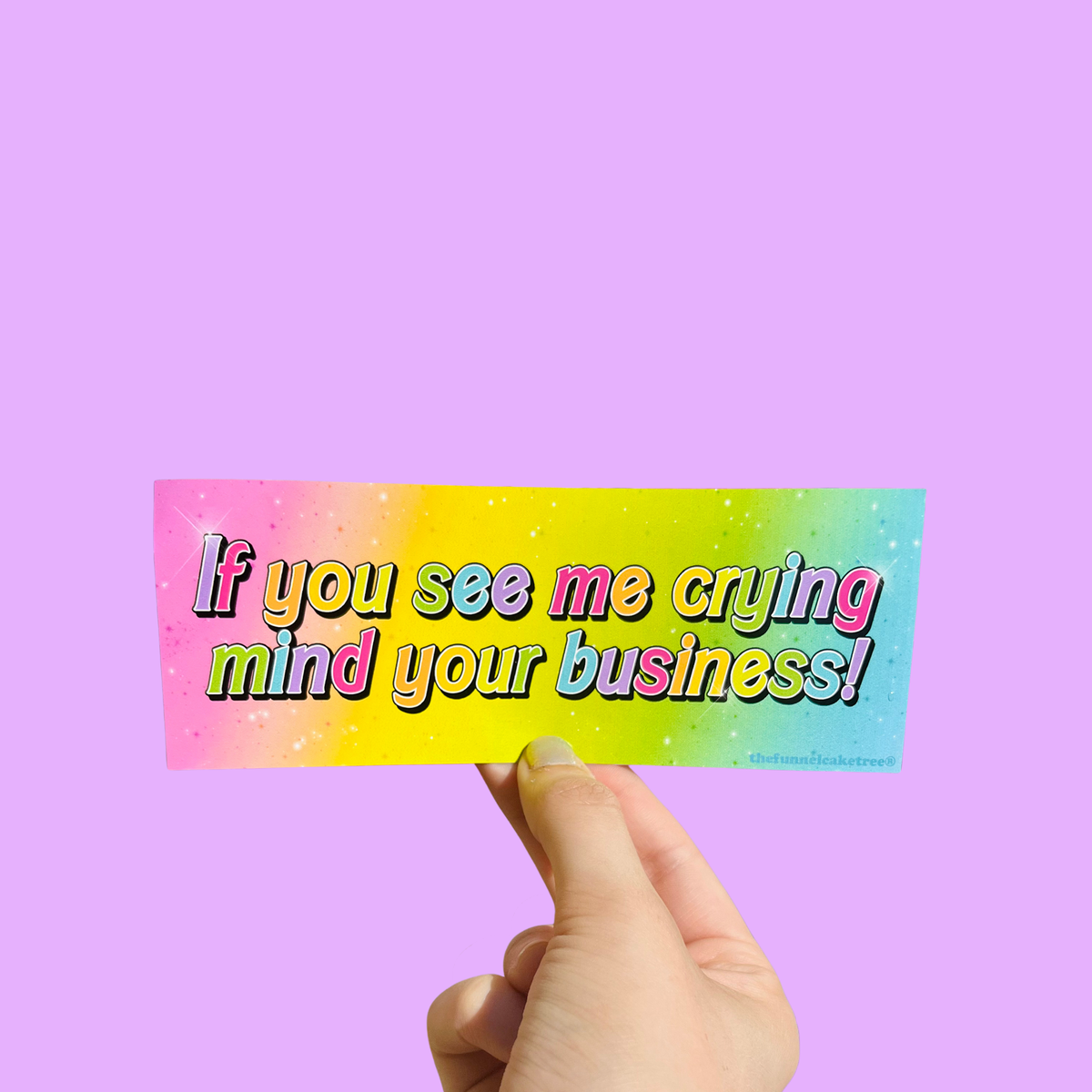 Hand holding a mini bumper sticker reading "if you see me crying mind your business." On a solid purple background.