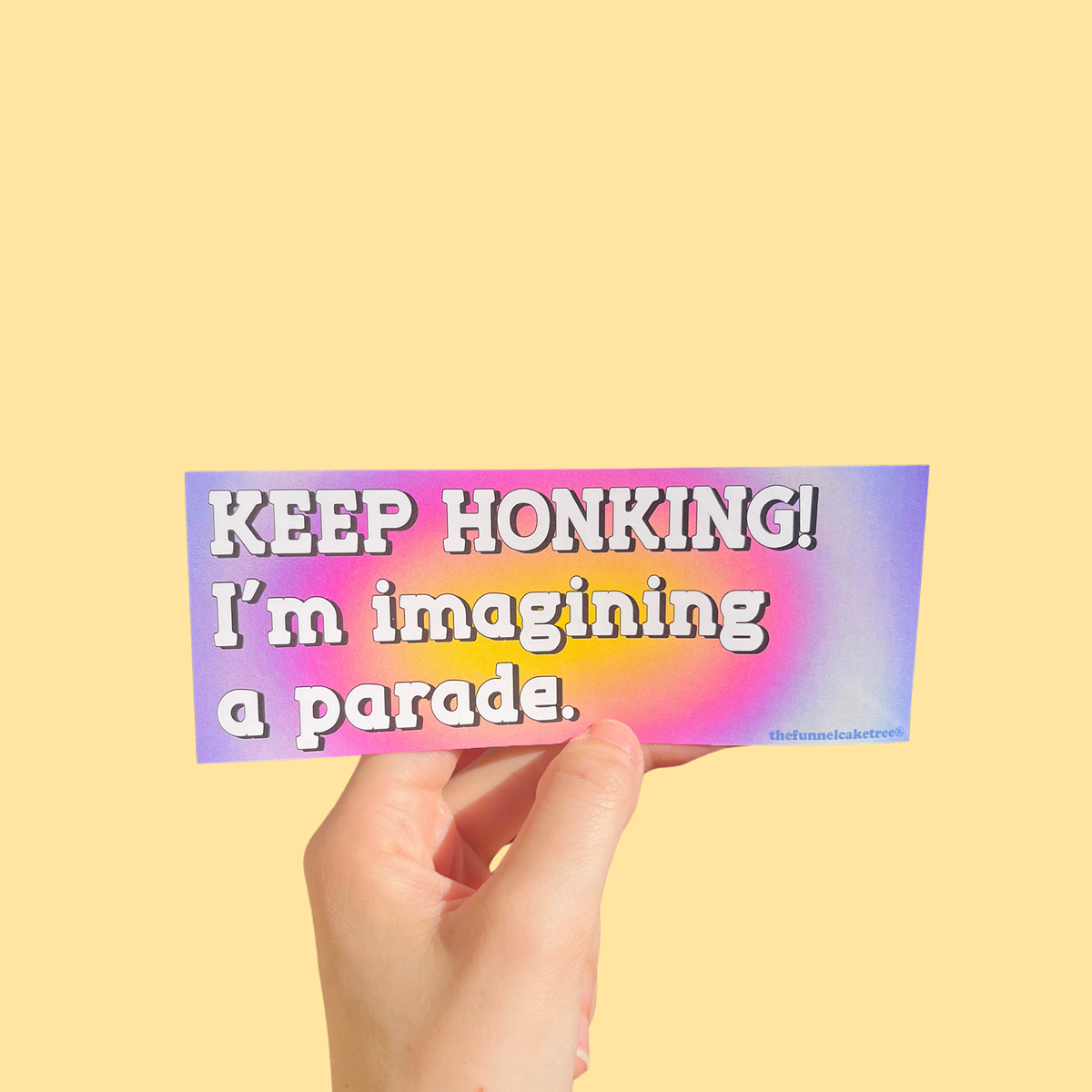 Hand holding a mini bumper sticker reading "keep honking im imagining a parade." On a solid yellow background.
