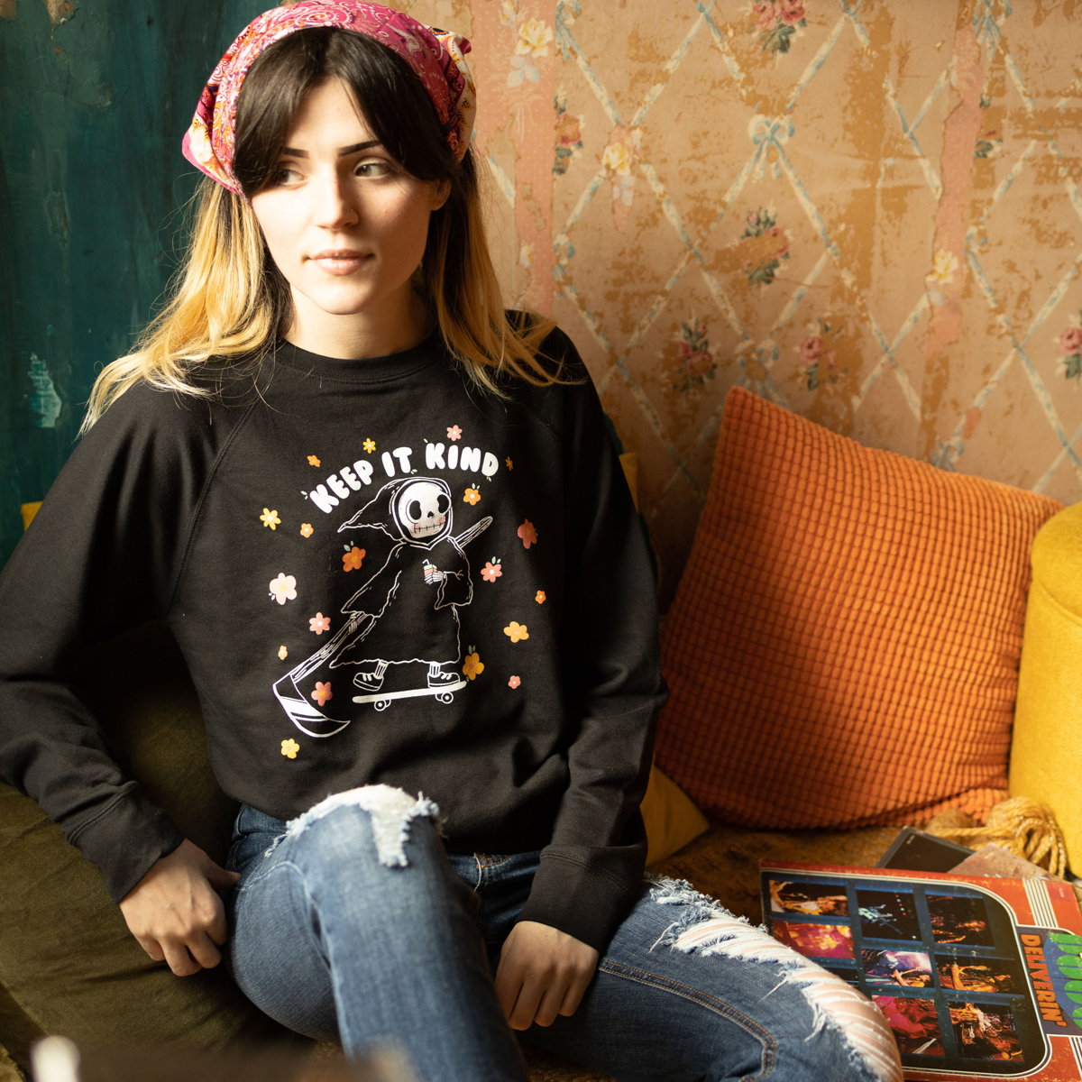 Izzy from The funnel cake tree sitting on a couch wearing a black sweatshirt with the keep it kind design in a room with a colorful wall. and 70s vibe