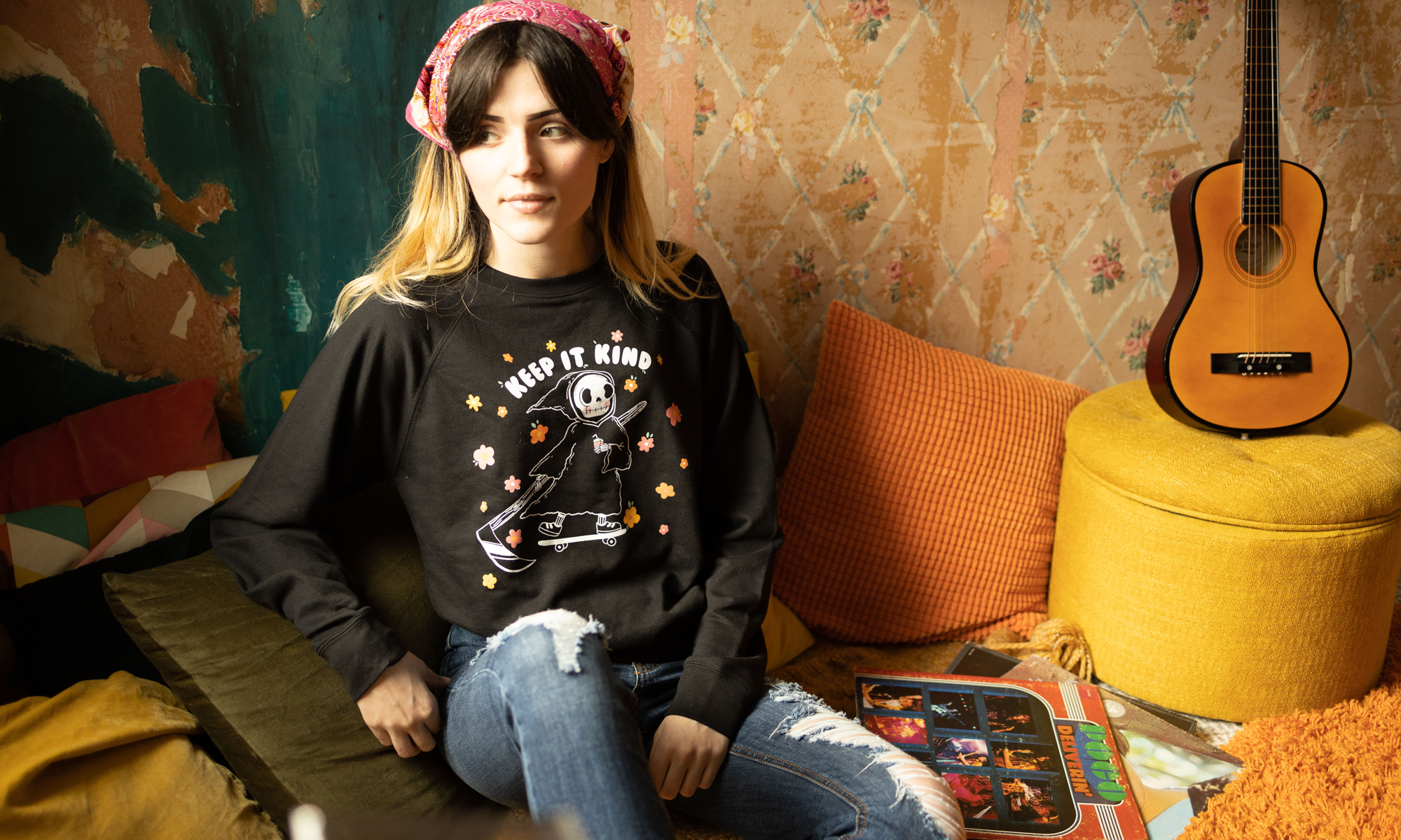 Izzy from The Funnel Cake Tree sitting on a couch wearing a black sweatshirt with her hand drawn keep it kind design in a room with a guitar and colorful wall.