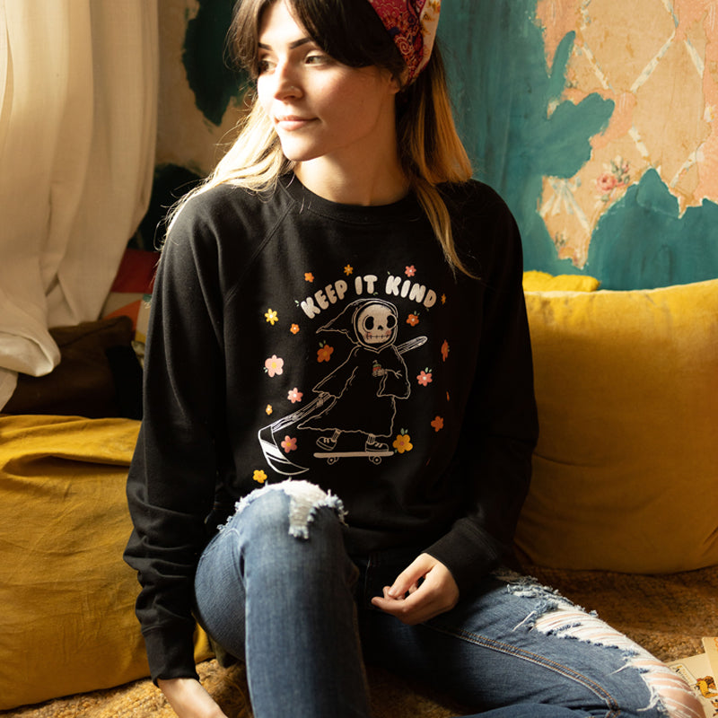 Izzy from The Funnel Cake Tree wearing a her art keep it kind on a black sweatshirt  sitting on a yellow pillow in a room with a 70s vibe