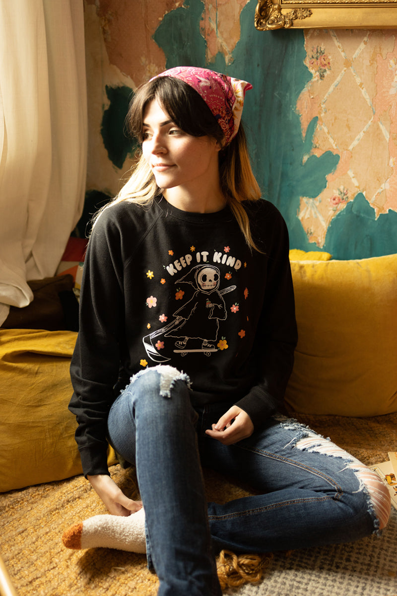 Izzy from The Funnel Cake Tree wearing a her art keep it kind on a black sweatshirt  sitting on a yellow pillow in a room with a 70s vibe