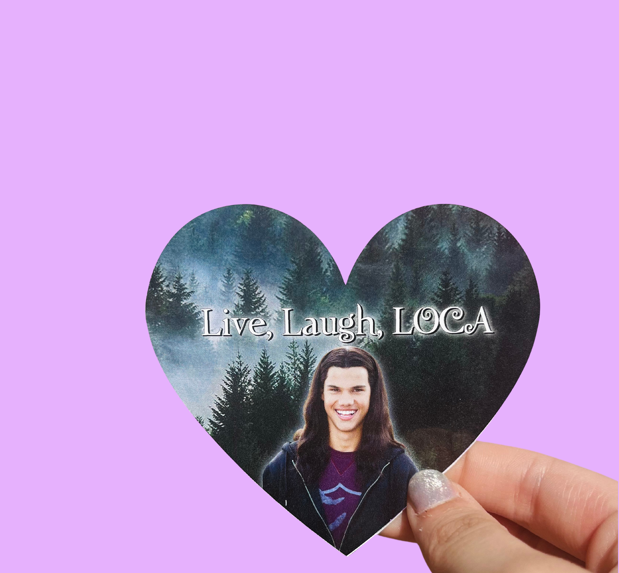 Hand holding a heart-shaped 'Live, Laugh, LOCA' sticker with a long-haired man's portrait and a forest background On a solid purple background.