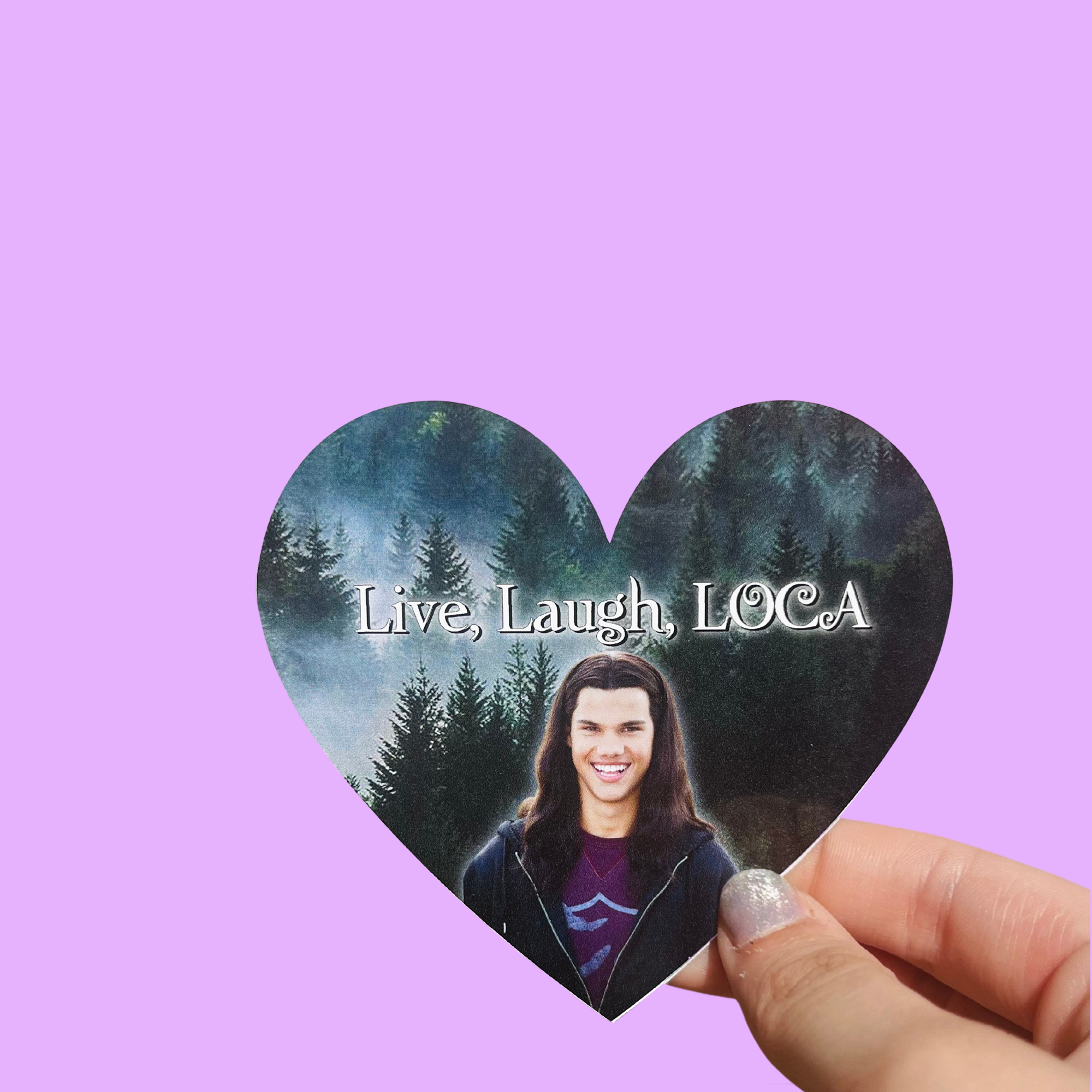 Hand holding a heart-shaped 'Live, Laugh, LOCA' sticker with a long-haired man's portrait and a forest background On a solid purple background.