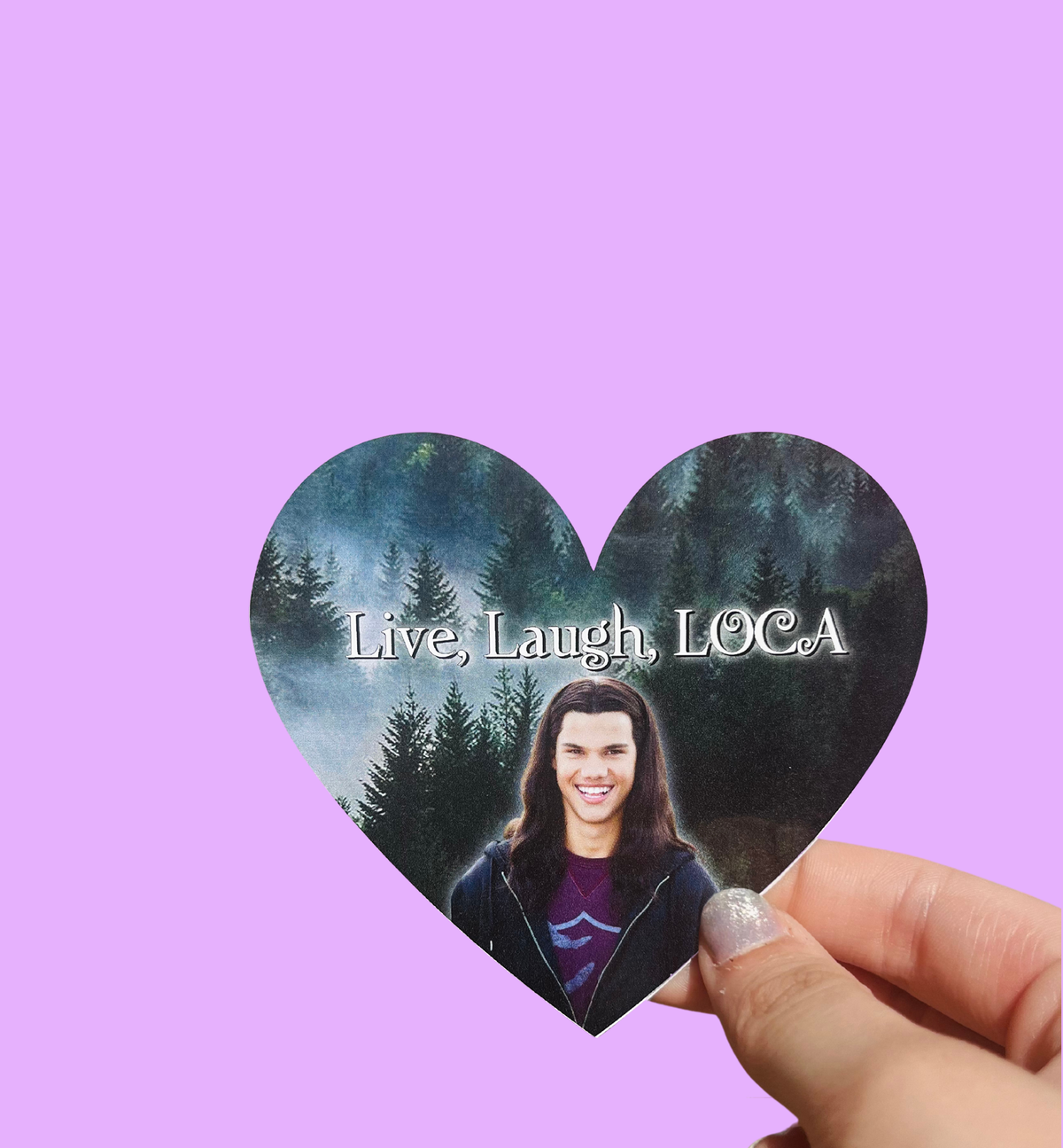 Hand holding a heart-shaped 'Live, Laugh, LOCA' sticker with a long-haired man's portrait and a forest background On a solid purple background.