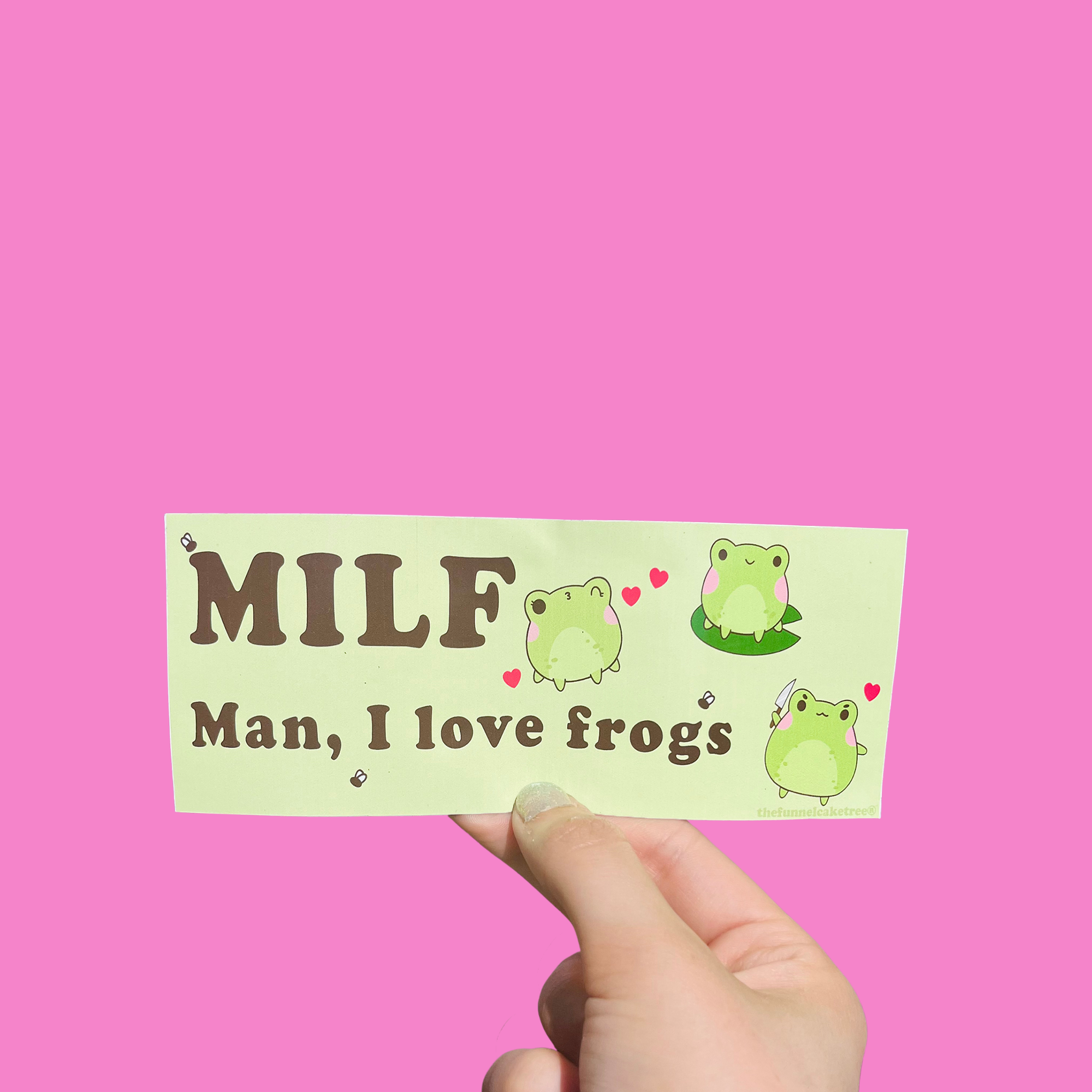 Hand holding a green mini bumper sticker reading 'MILF — Man, I love frogs' with cartoon frogs and hearts On a solid pink background.