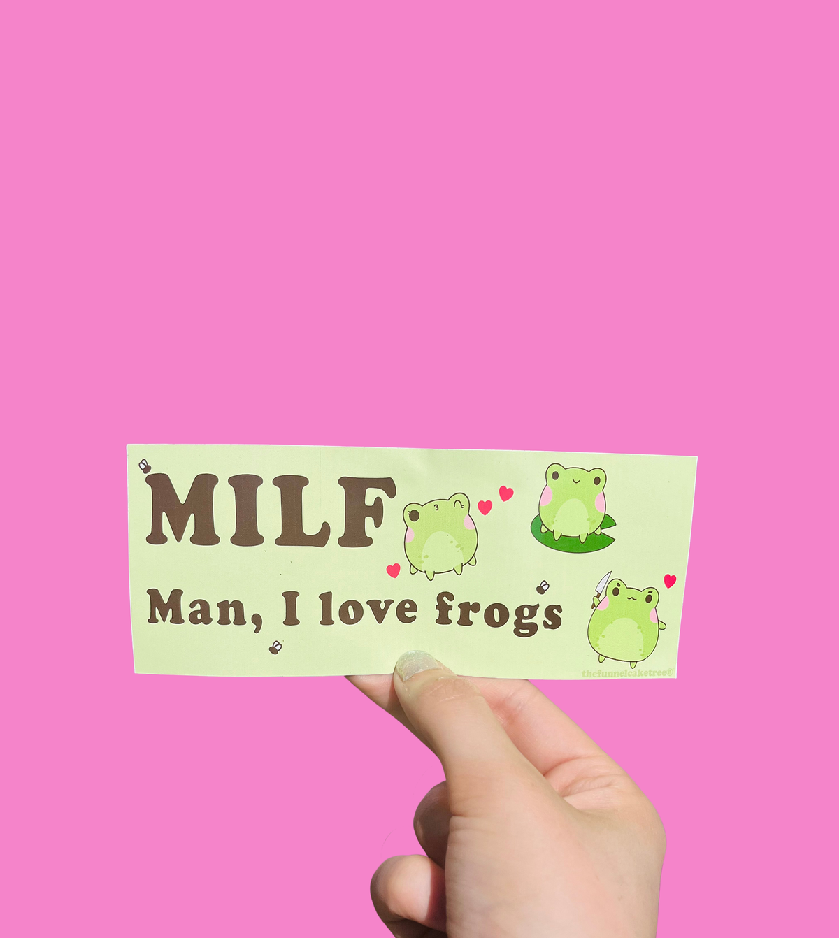 Hand holding a green mini bumper sticker reading 'MILF — Man, I love frogs' with cartoon frogs and hearts On a solid pink background.