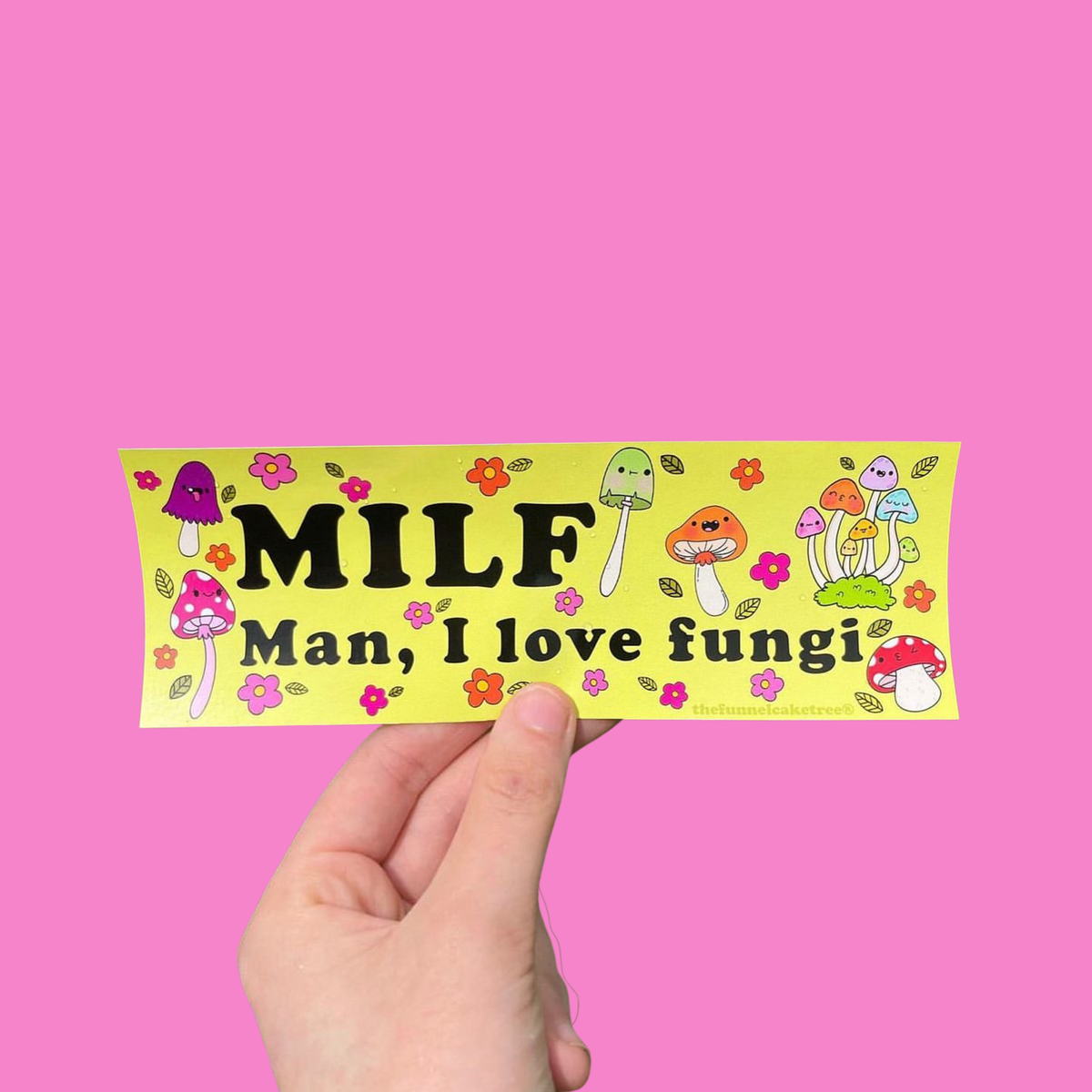 Hand holding a yellow-green 'MILF — Man, I love fungi' mini bumper sticker with colorful mushrooms and flowers On a solid pink background.