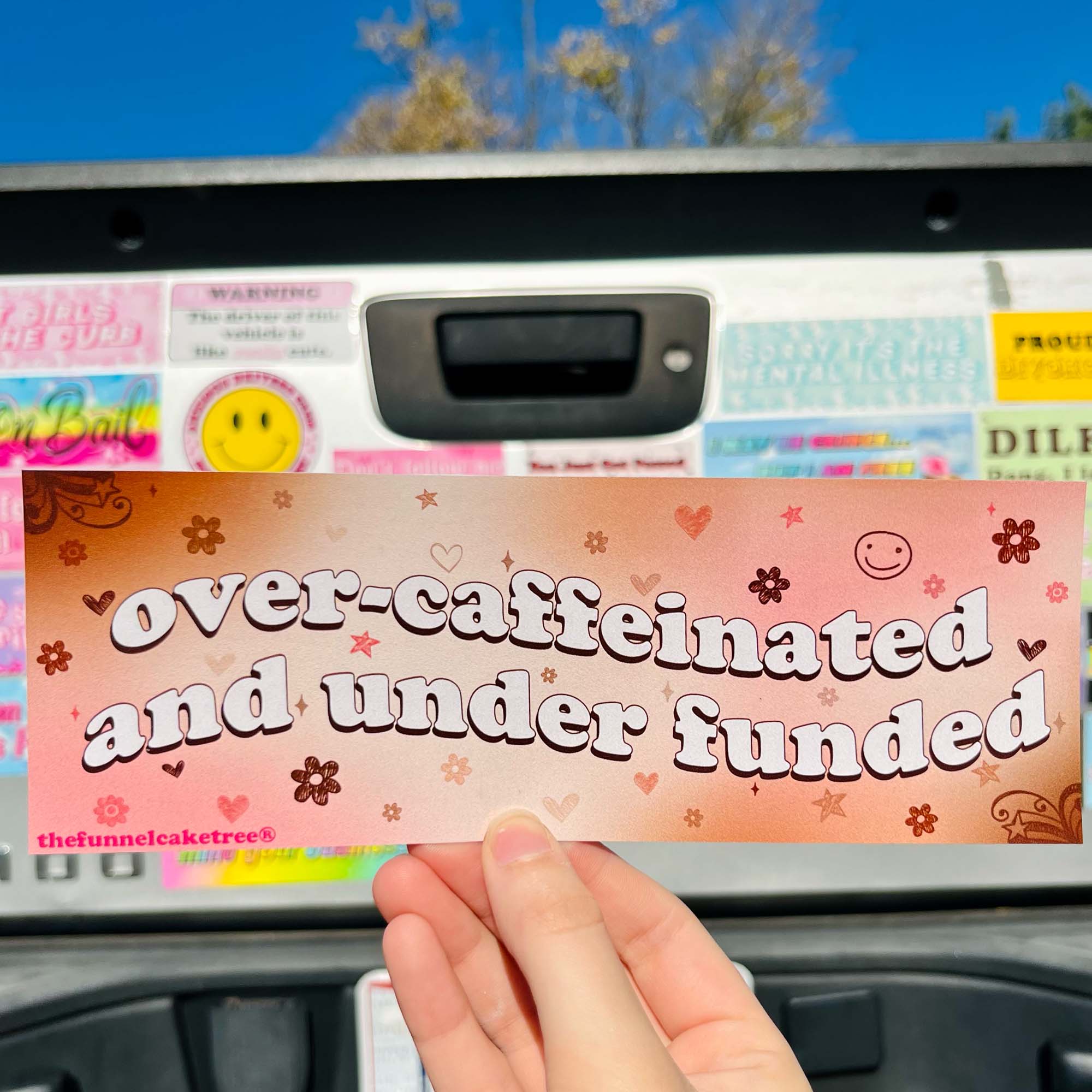 A hand holding a bumper sticker with the text 'over-caféinated and underfunded' on it, with a pink background and decorative elements.