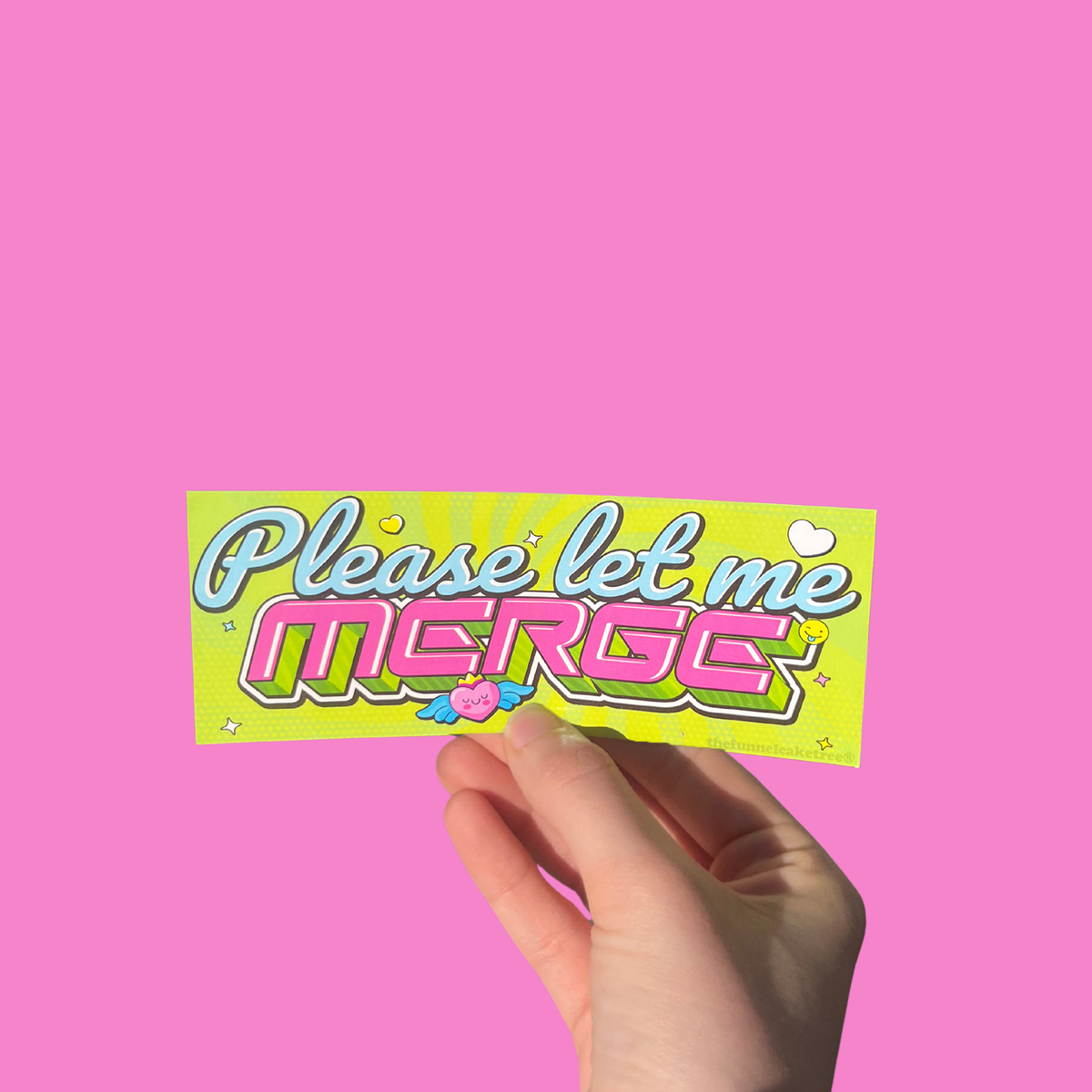 Hand holding a lime green Y2K-style mini bumper sticker reading 'Please let me MERGE' in pink and white letters On a solid pink background.