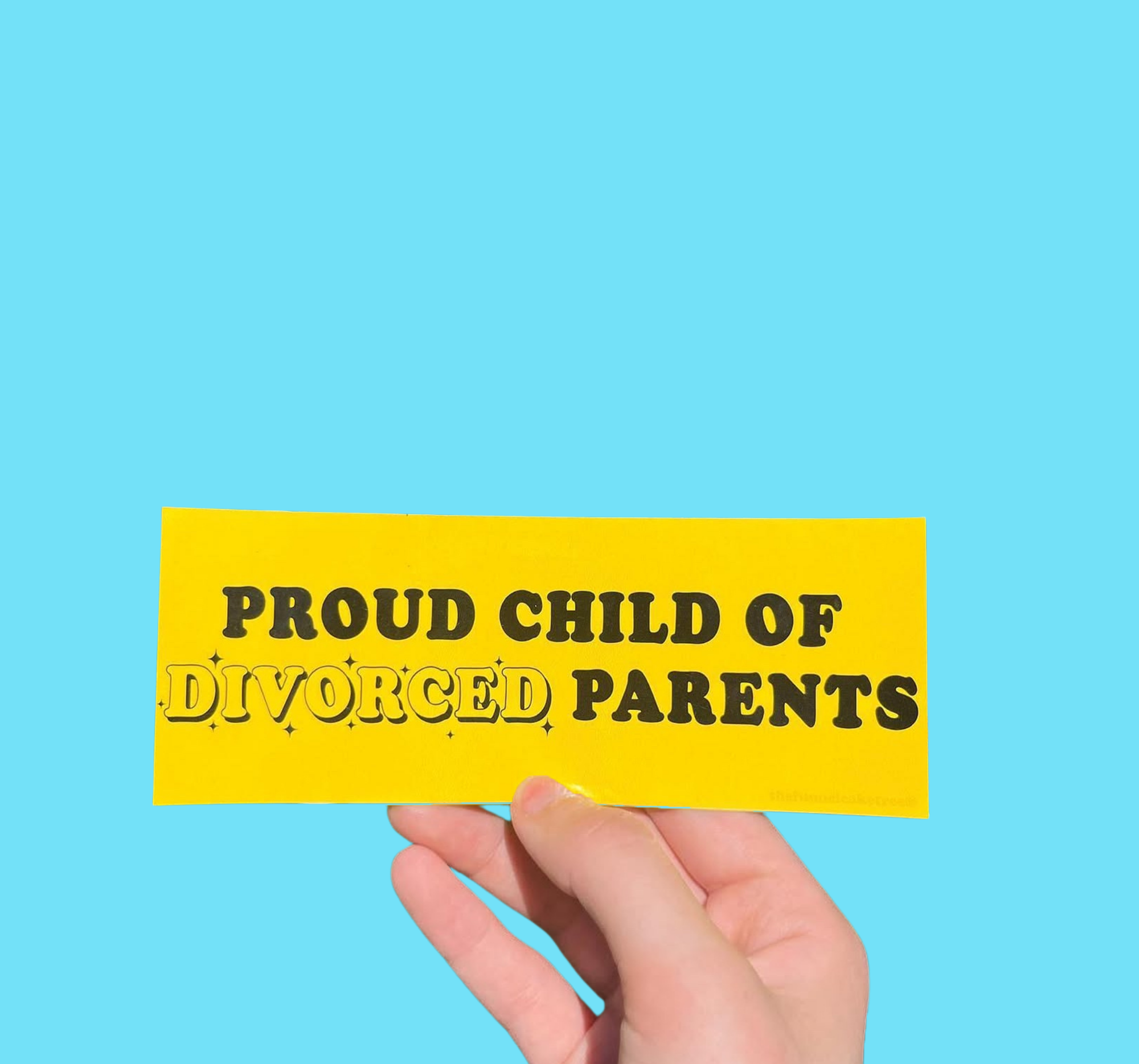 Hand holding a yellow mini bumper sticker reading 'Proud child of divorced parents' in black lettering On a solid teal background.