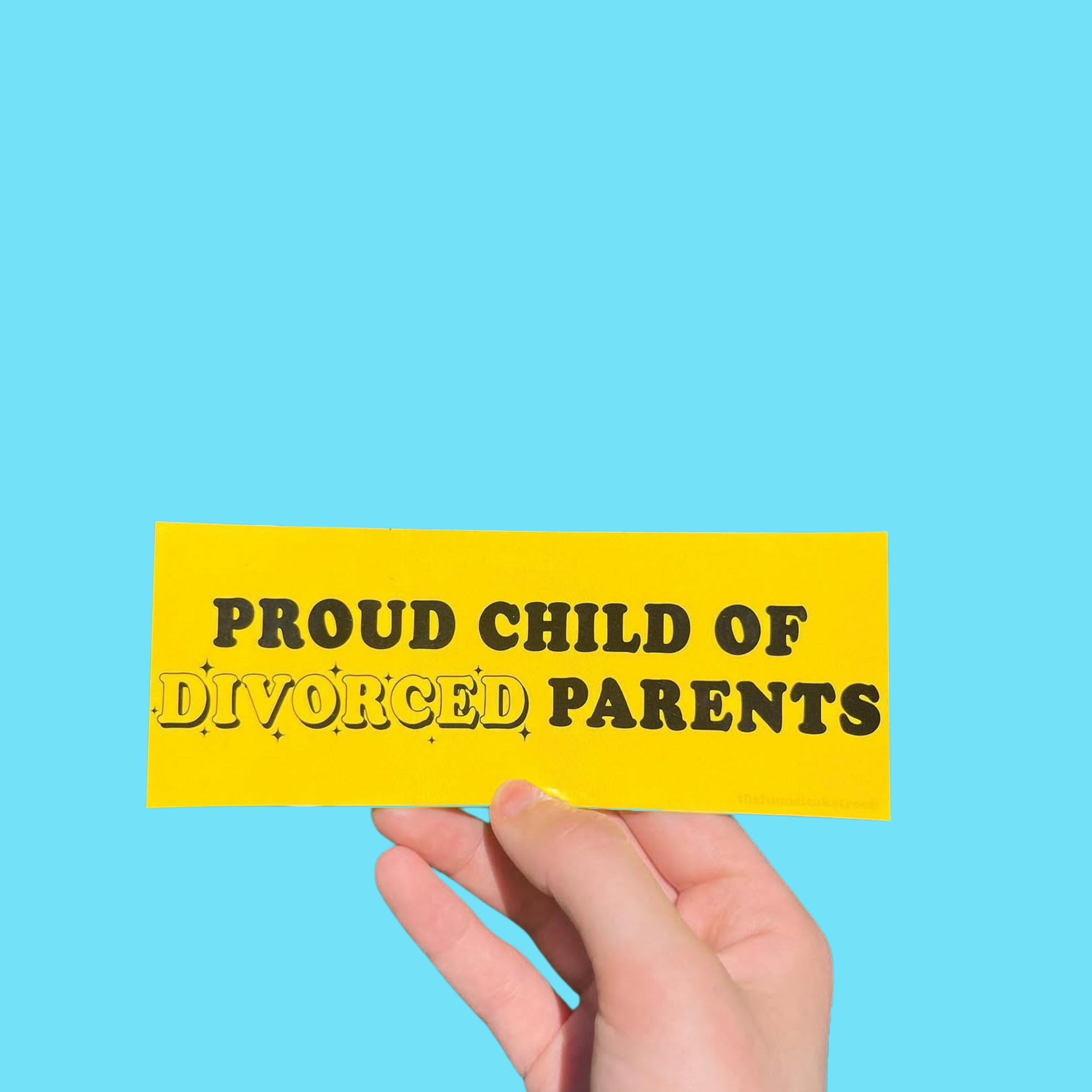 Hand holding a yellow mini bumper sticker reading 'Proud child of divorced parents' in black lettering On a solid teal background.