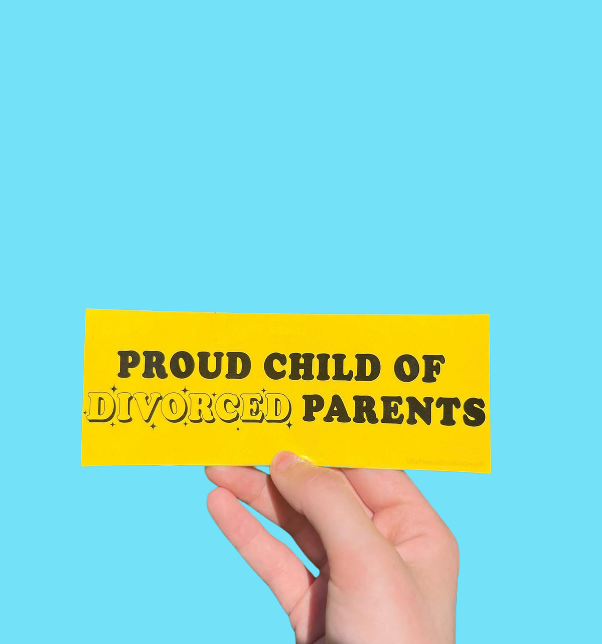 Hand holding a yellow mini bumper sticker reading 'Proud child of divorced parents' in black lettering On a solid teal background.