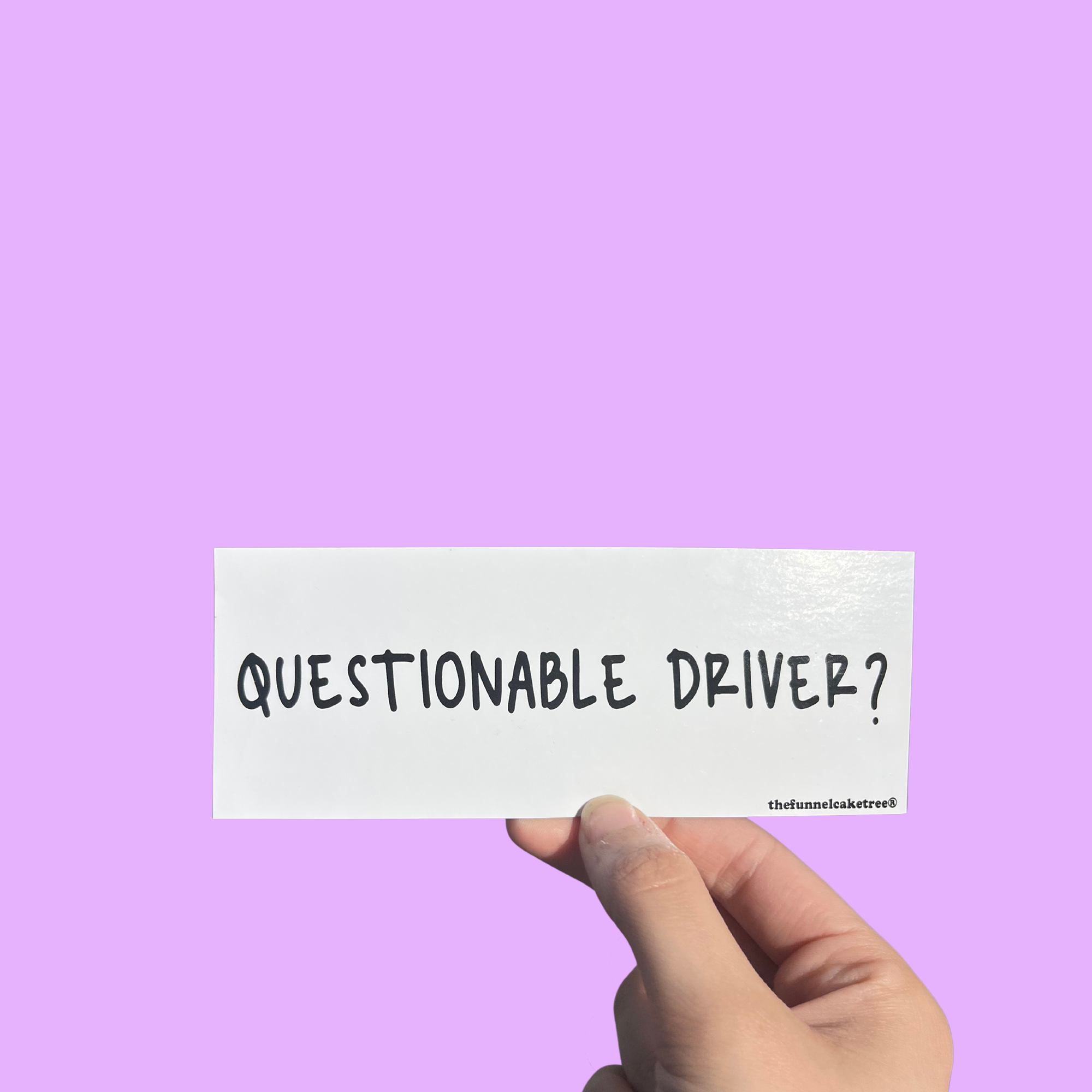 Hand holding a mini bumper sticker reading "questionable driver." On a solid purple background.