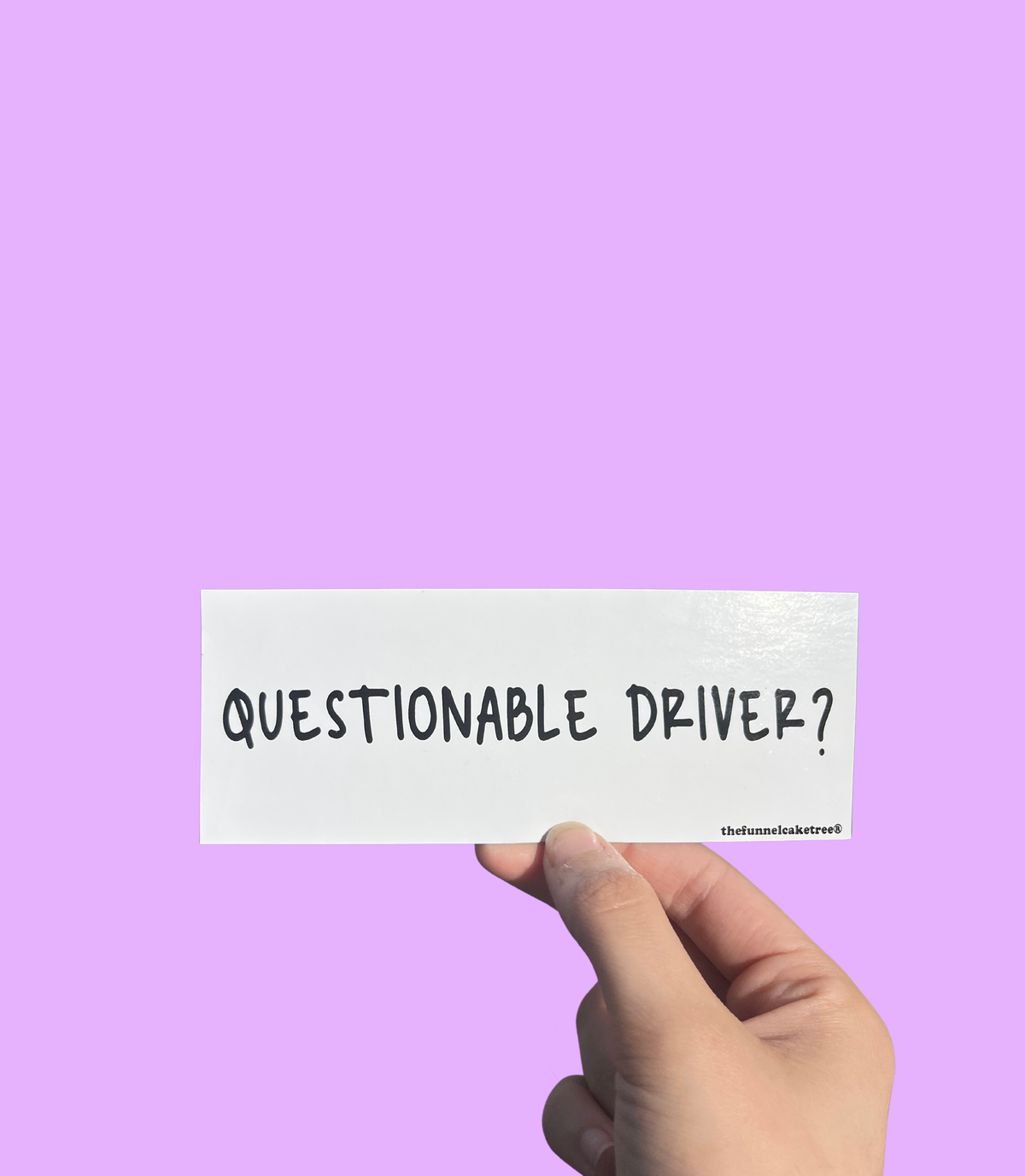 Hand holding a mini bumper sticker reading "questionable driver." On a solid purple background.