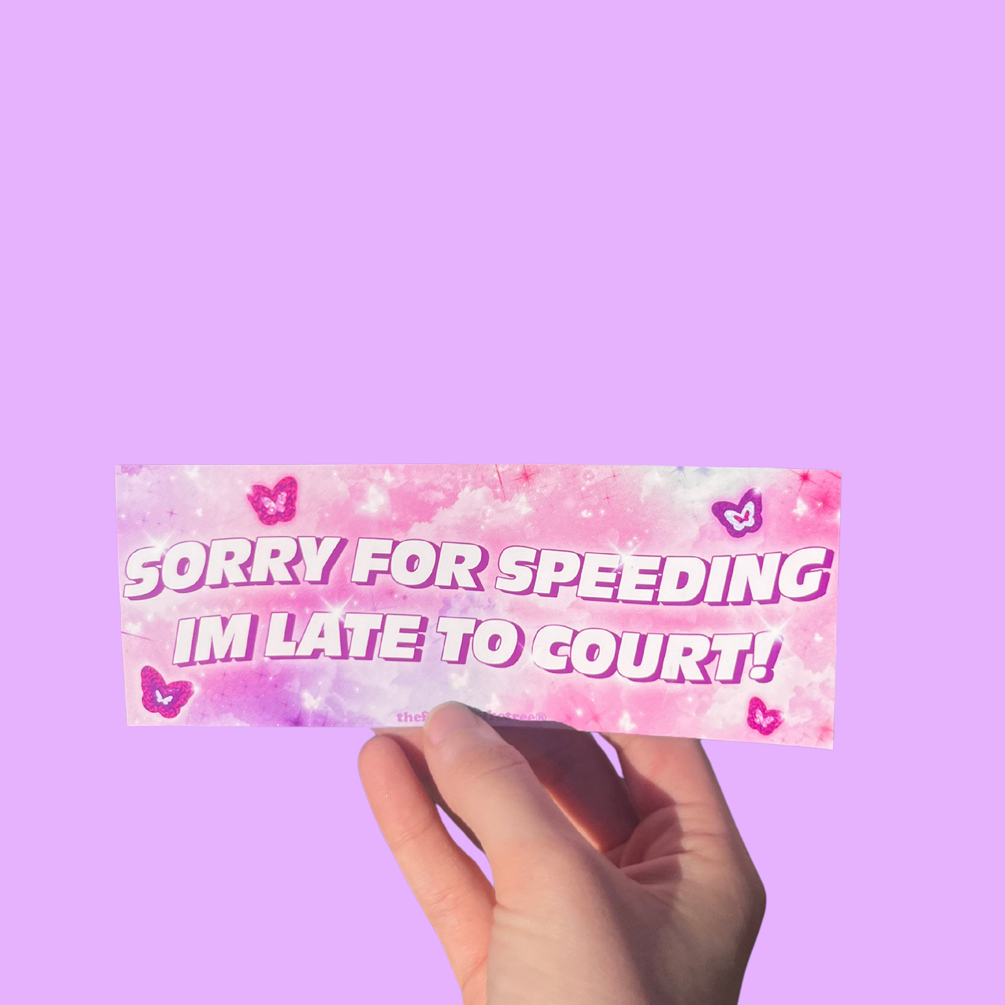 Hand holding a pink mini bumper sticker reading 'Sorry for speeding I'm late to court!' with hearts On a solid purple background.