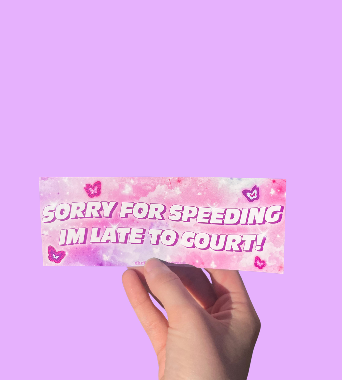 Hand holding a pink mini bumper sticker reading 'Sorry for speeding I'm late to court!' with hearts On a solid purple background.