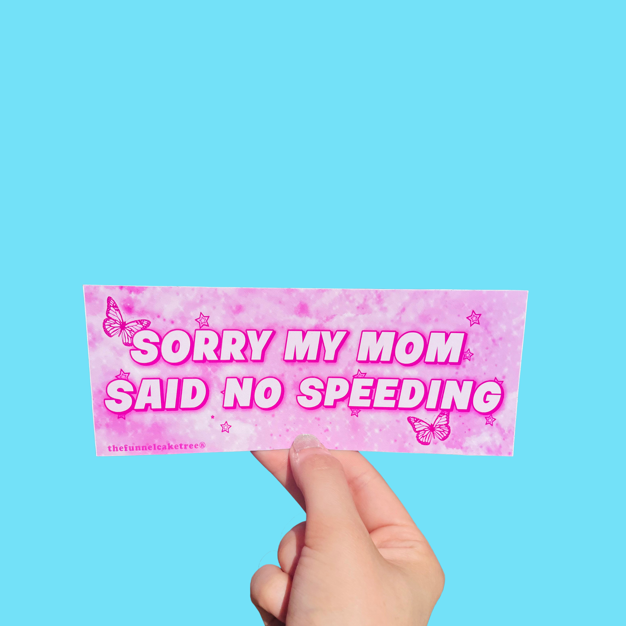 Hand holding a hot-pink mini bumper sticker reading 'Sorry my mom said no speeding' with butterflies and stars On a solid teal background.