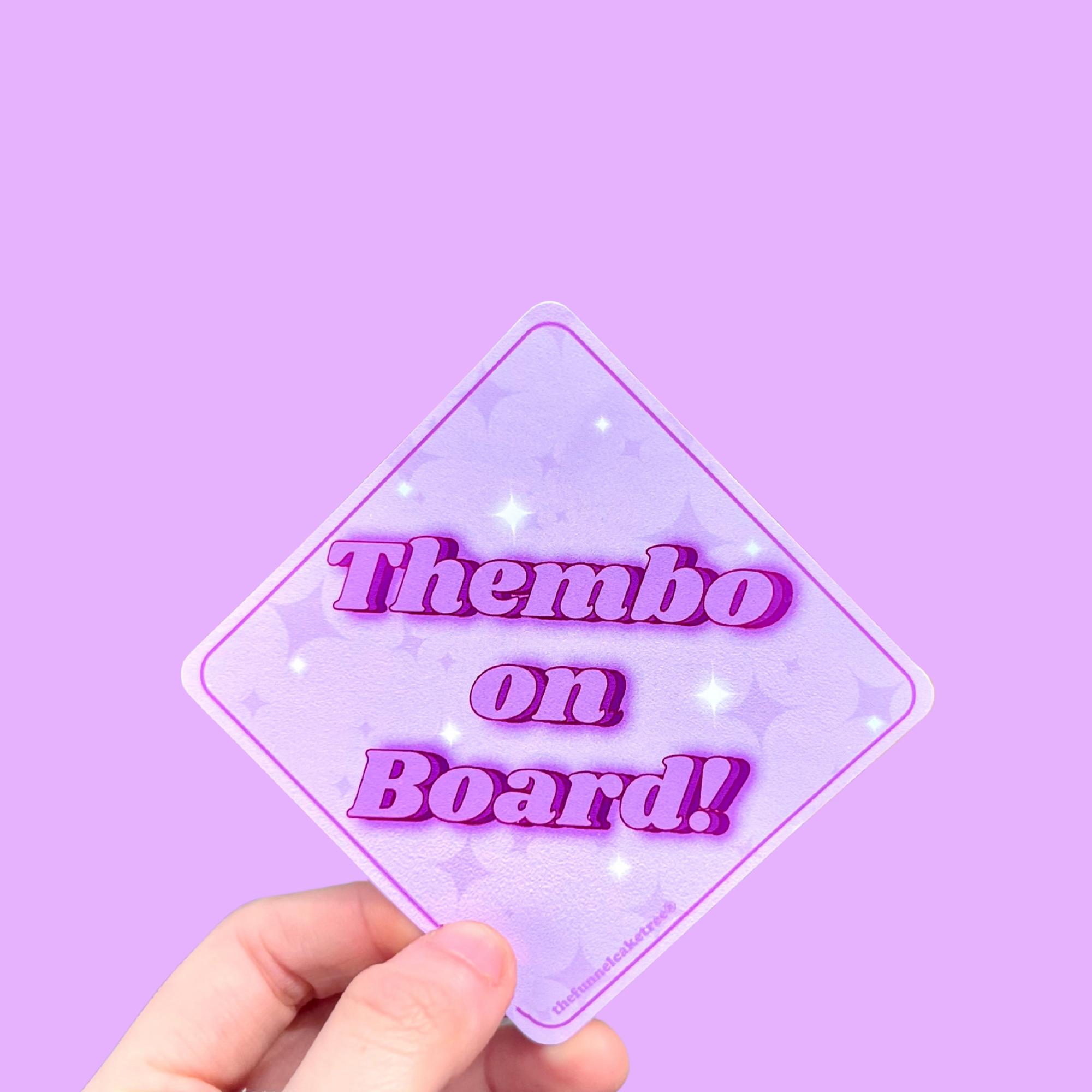 Hand holding a mini bumper sticker reading "thembo on board." On a solid purple background.