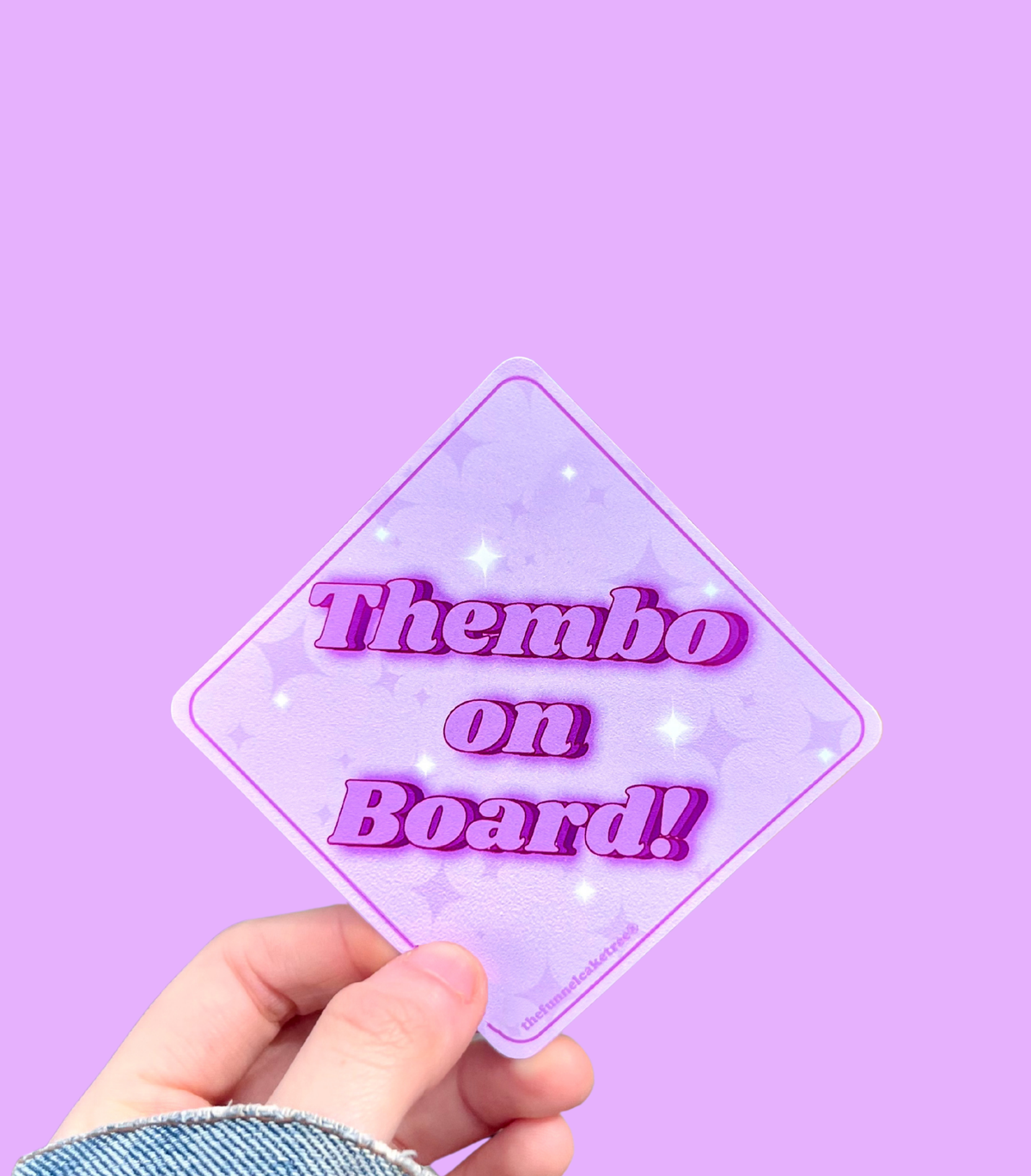 Hand holding a mini bumper sticker reading "thembo on board." On a solid purple background.