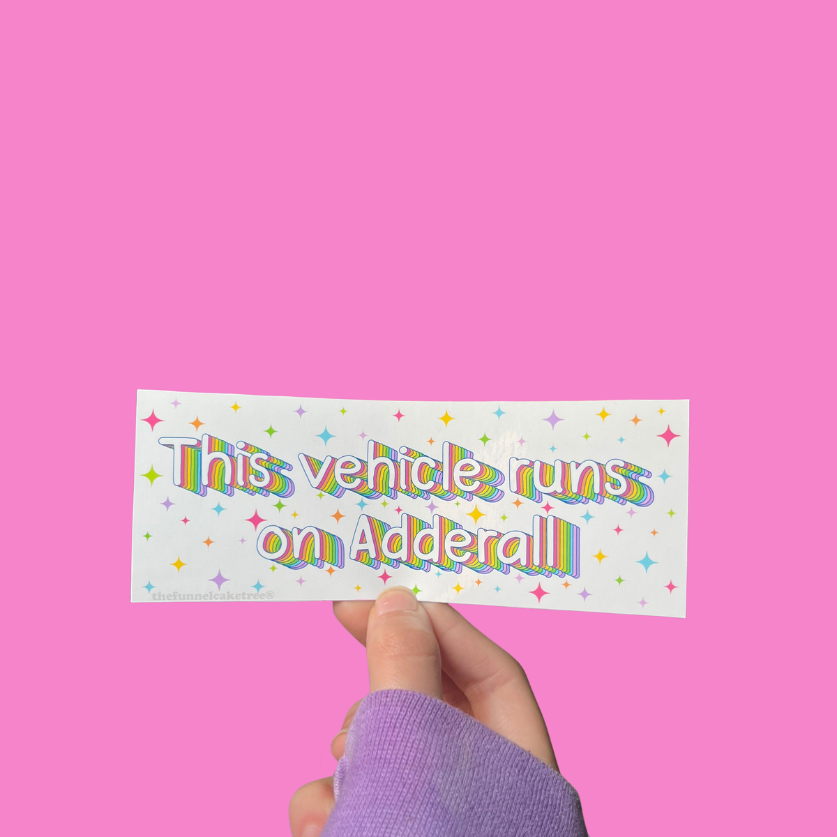 Hand holding a mini bumper sticker reading "this vehicle runs on adderall." On a solid pink background.