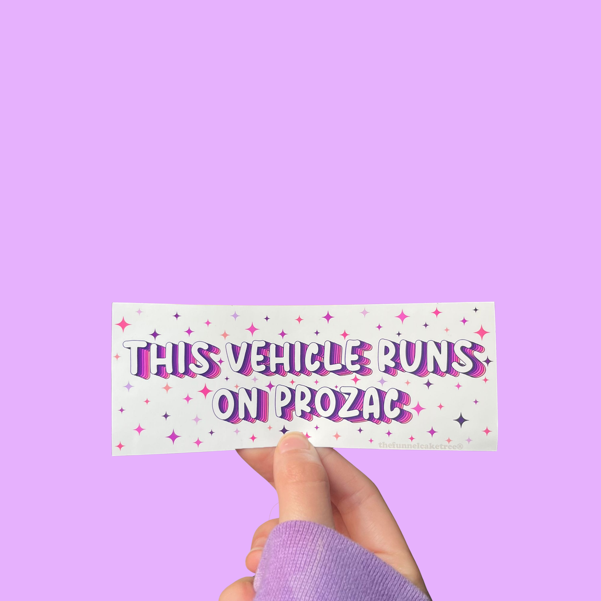 Hand holding a mini bumper sticker reading "this vehicle runs on prozac." On a solid purple background.