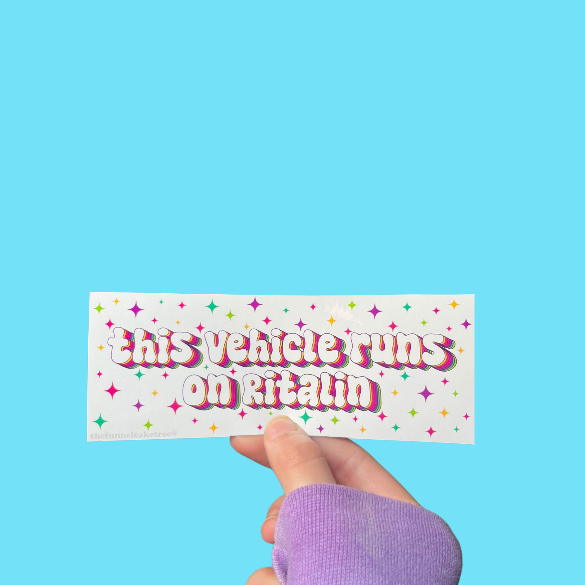 Hand holding a mini bumper sticker reading "this vehicle runs on ritalin." On a solid teal background.
