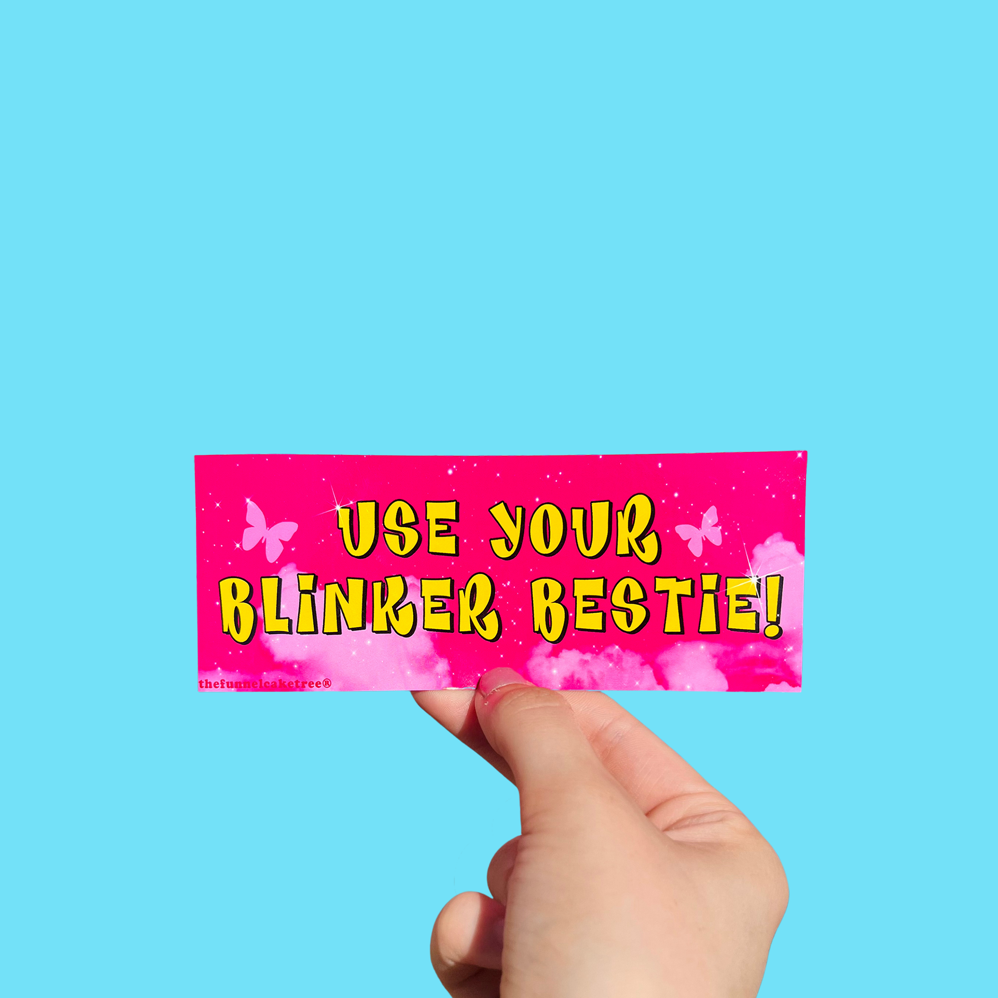 Hand holding a mini bumper sticker reading "use your blinker bestie." On a solid teal background.