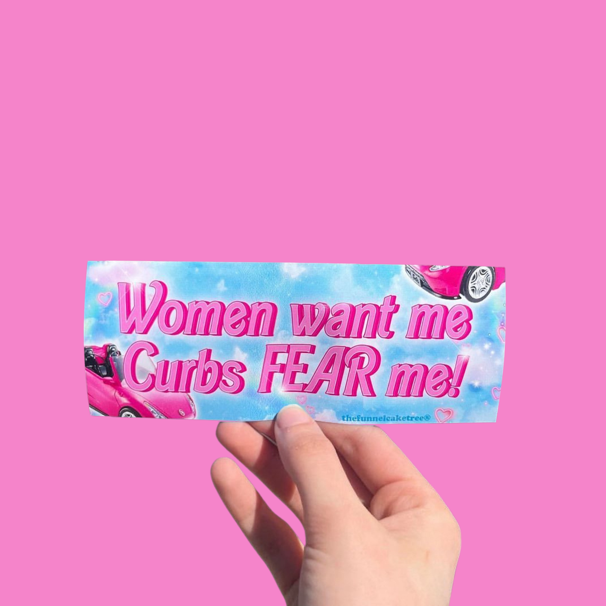 Hand holding a mini bumper sticker reading "women want me curbs fear me." On a solid pink background.