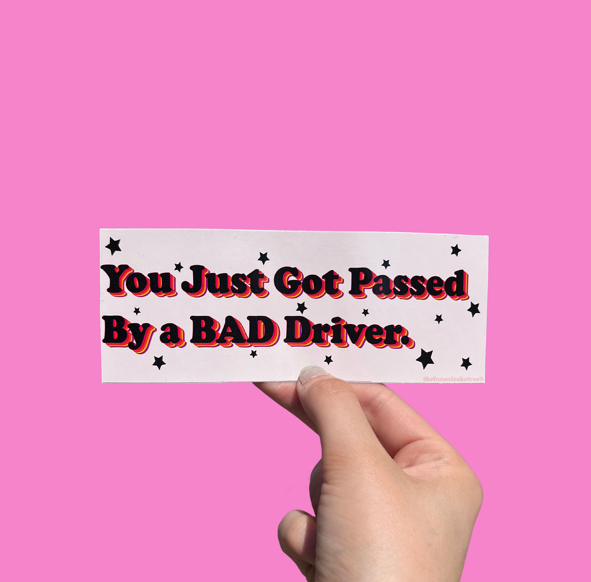 Hand holding a mini bumper sticker reading "you just got passed by a bad driver." On a solid pink background.