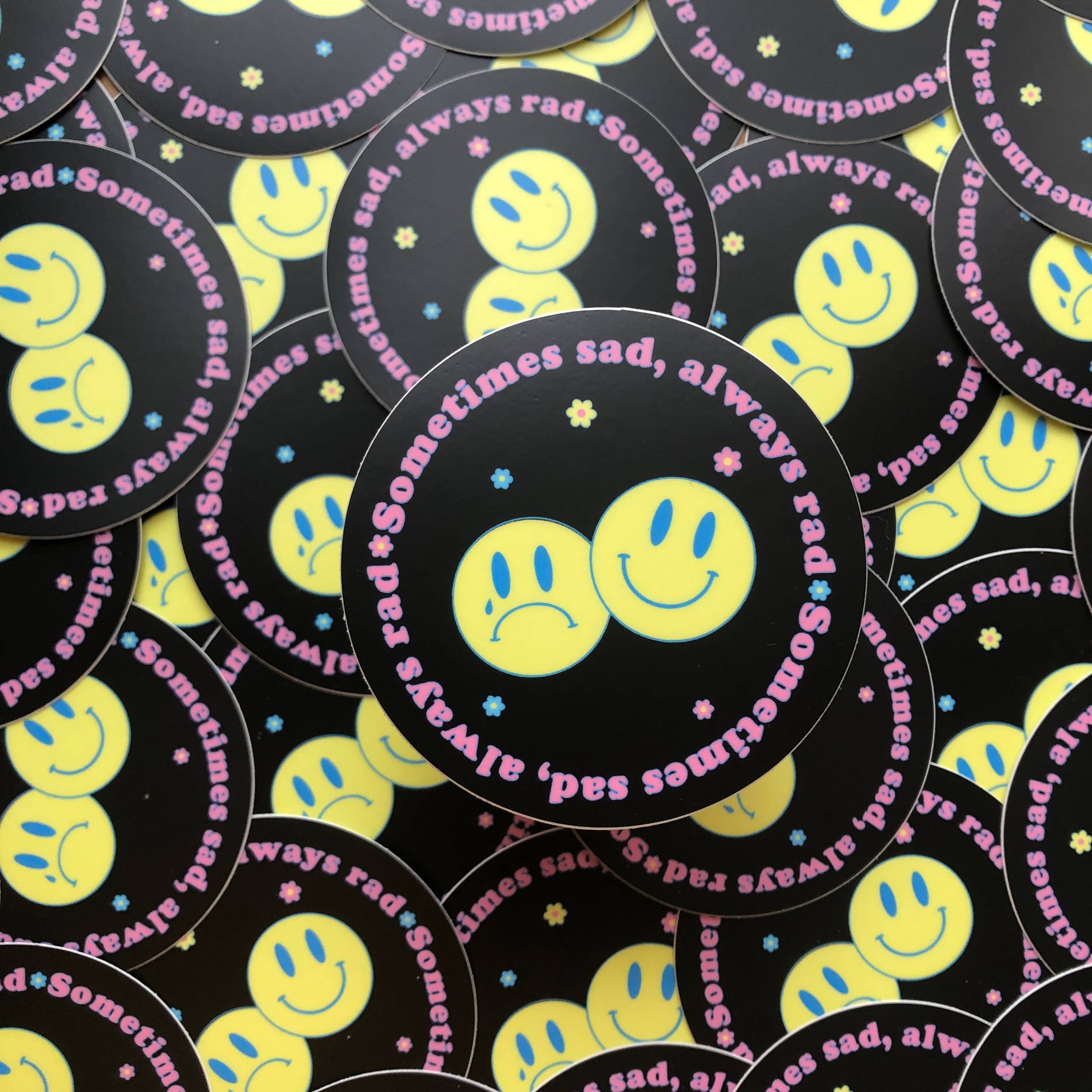 Pile of round black stickers with yellow smiley faces and pink text reading Sometimes sad, always rad.