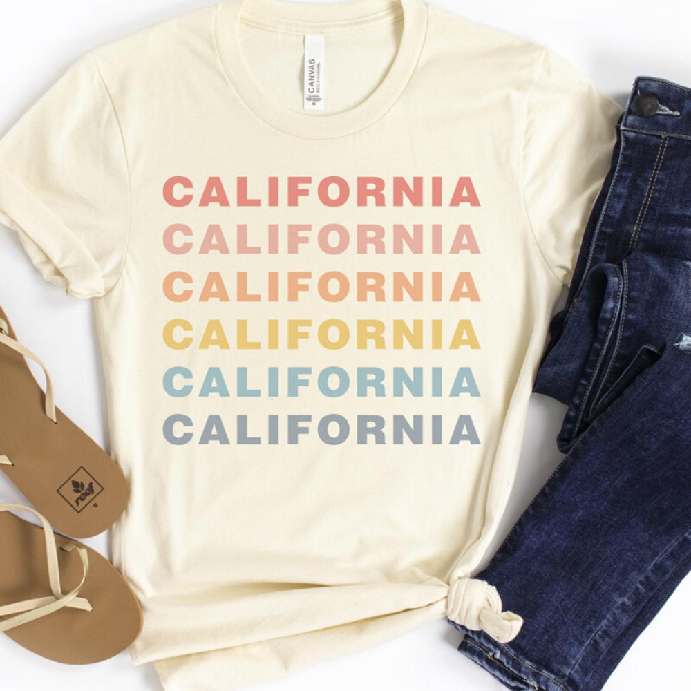 Cream-colored unisex t-shirt with repeating pastel text reading “CALIFORNIA” in coral, peach, yellow, teal, and blue, styled flat with jeans and sandals.