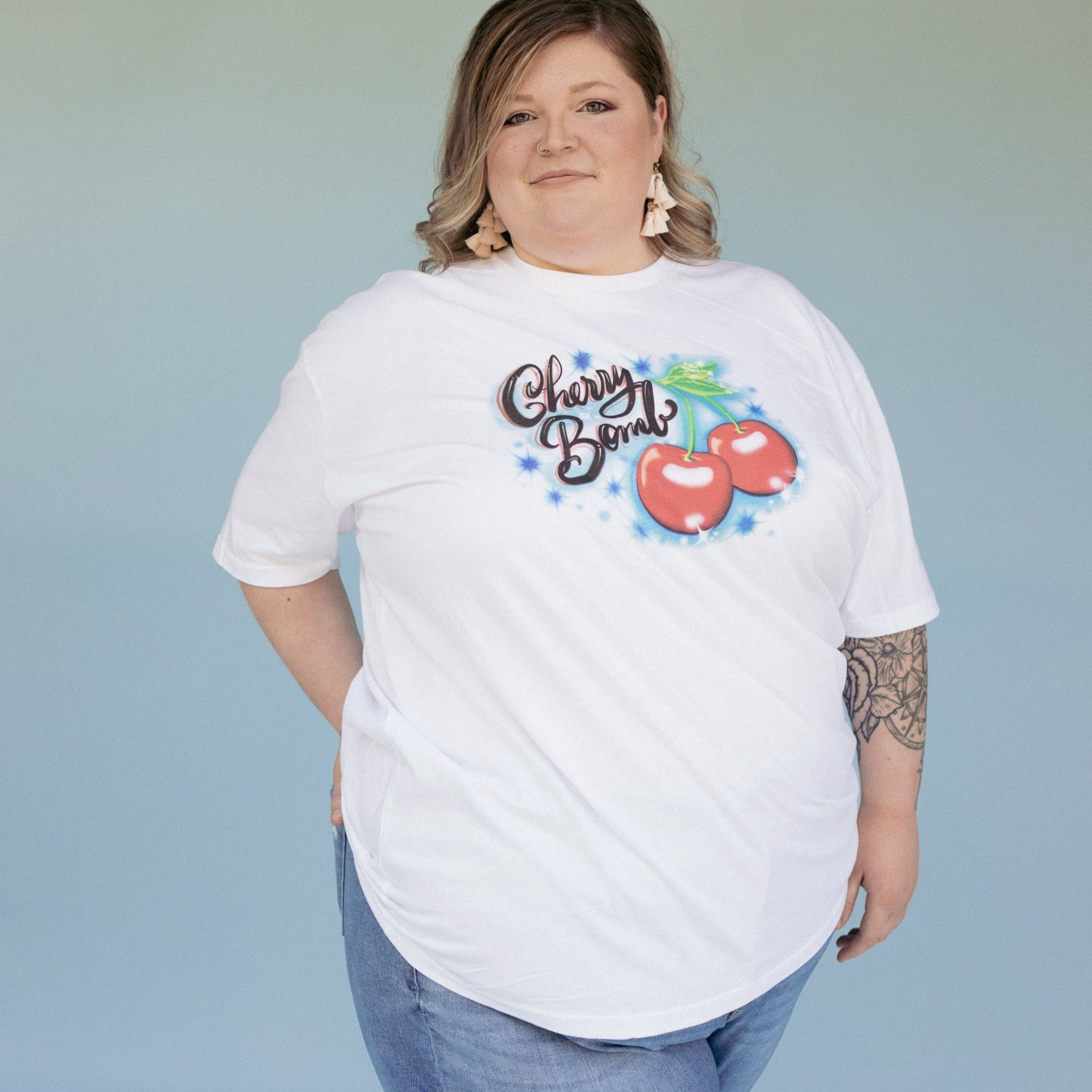 Model wearing The Funnel Cake Tree “Cherry Bomb” white graphic tee featuring bold red cherries and retro airbrush-style lettering on a soft unisex cotton shirt.