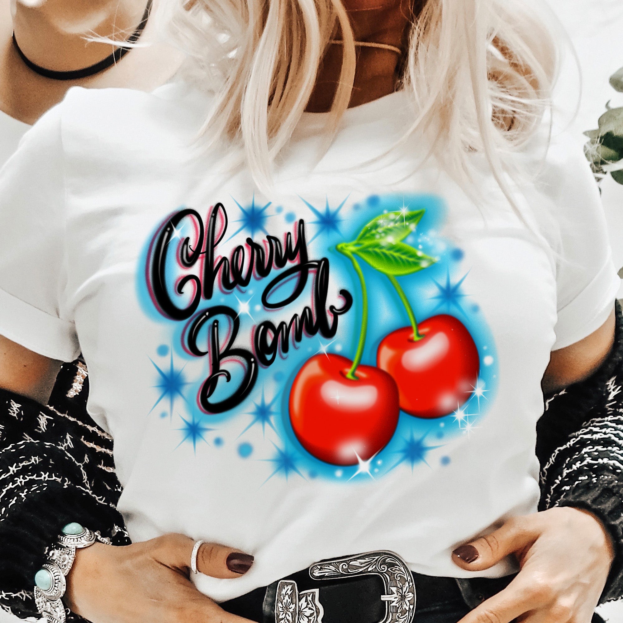 Close-up of The Funnel Cake Tree “Cherry Bomb” white graphic tee featuring bold red cherries, blue sparkle accents, and retro airbrush-style lettering for a vintage Y2K look.