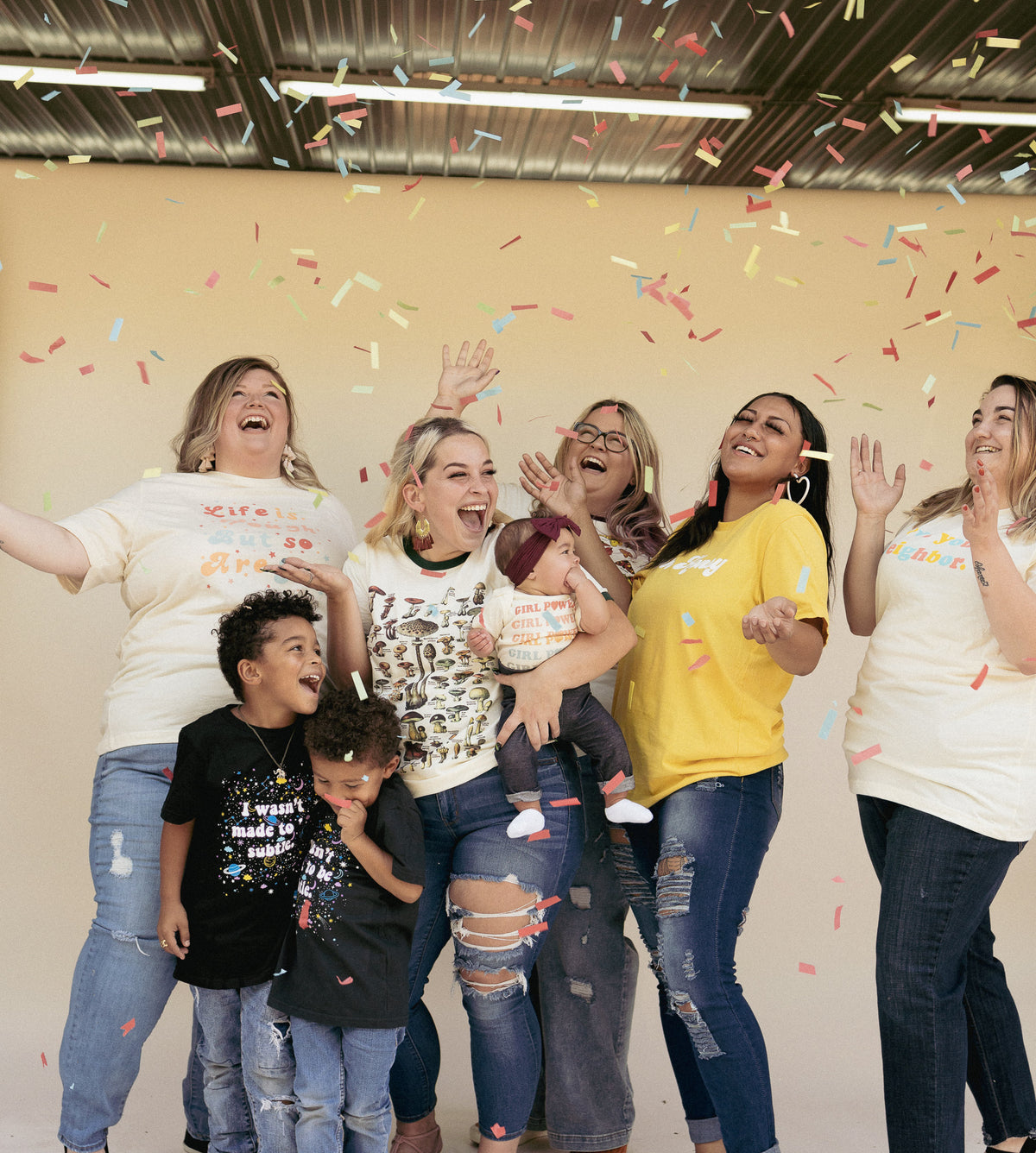 Group of women and children wearing colorful vintage-style graphic tees from The Funnel Cake Tree, celebrating under falling confetti and smiling showcasing mushroom, girl power, and retro-inspired designs.