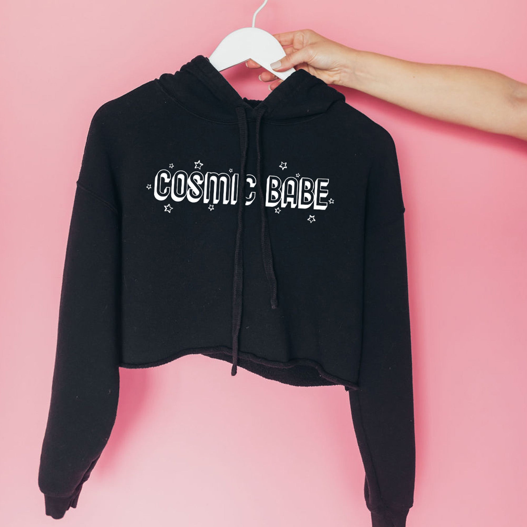 The Funnel Cake Tree “Cosmic Babe” raw-edge cropped black hoodie featuring bold white retro lettering and star details, displayed against a pink background for a stylish celestial look.