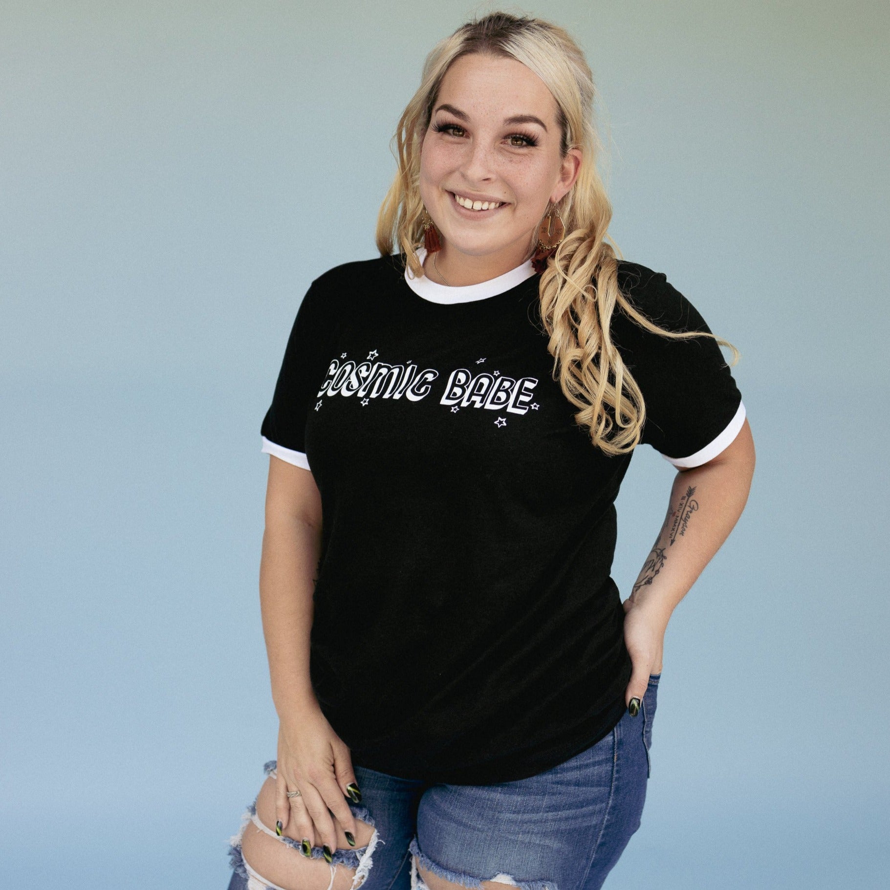 The Funnel Cake Tree “Cosmic Babe” black ringer tee with white trim, featuring bold retro star lettering. Plus-size model styled in casual ripped denim for a laid-back celestial look.
