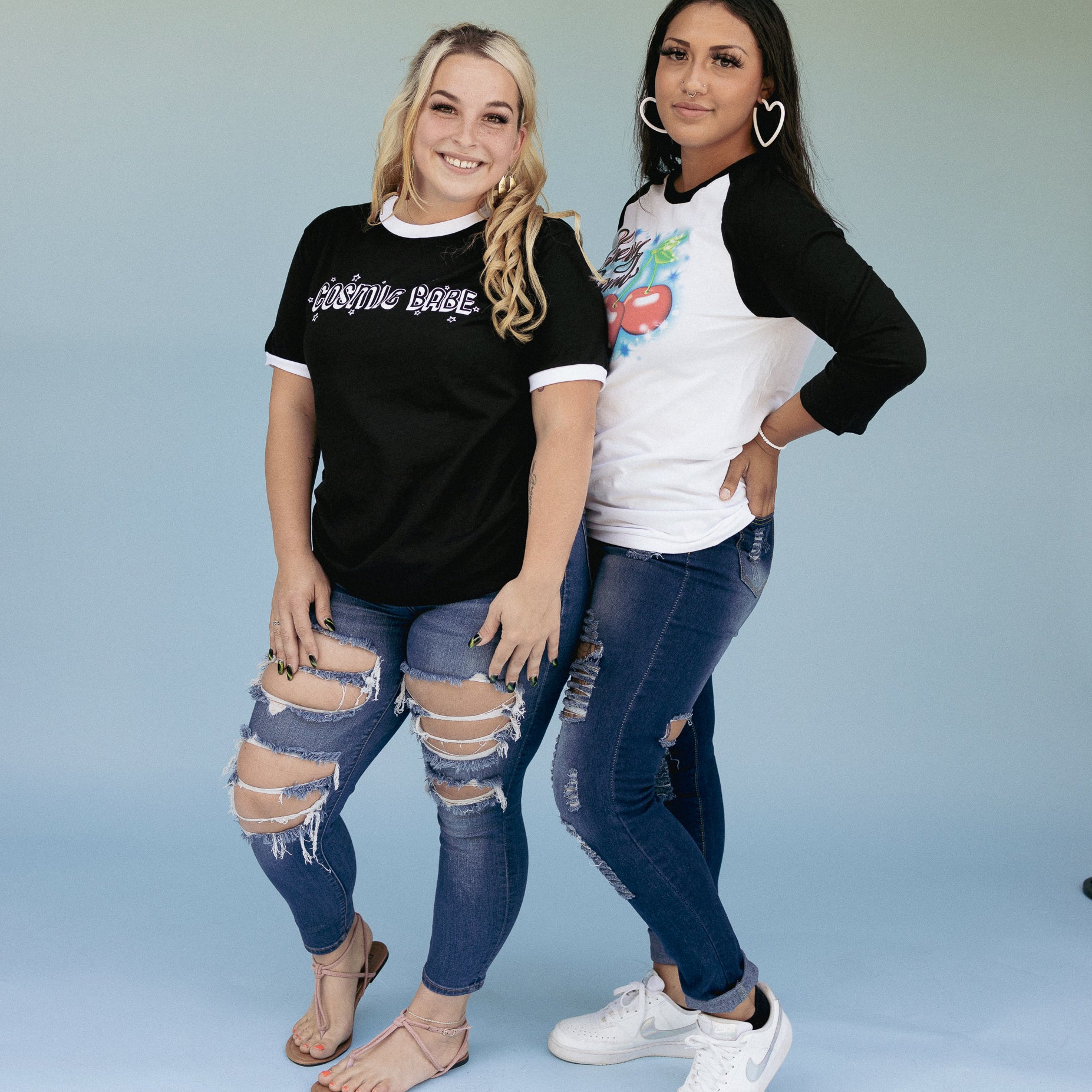 Two models wearing The Funnel Cake Tree graphic tees, one in the “Cosmic Babe” black ringer tee with white trim, and the other in the “Cherry Bomb” raglan tee with red cherry print styled with ripped jeans for a fun, retro-inspired look.