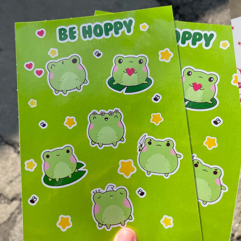 Two green cards with cartoon frogs and 'BE HOPPY' text on a stone surface.
