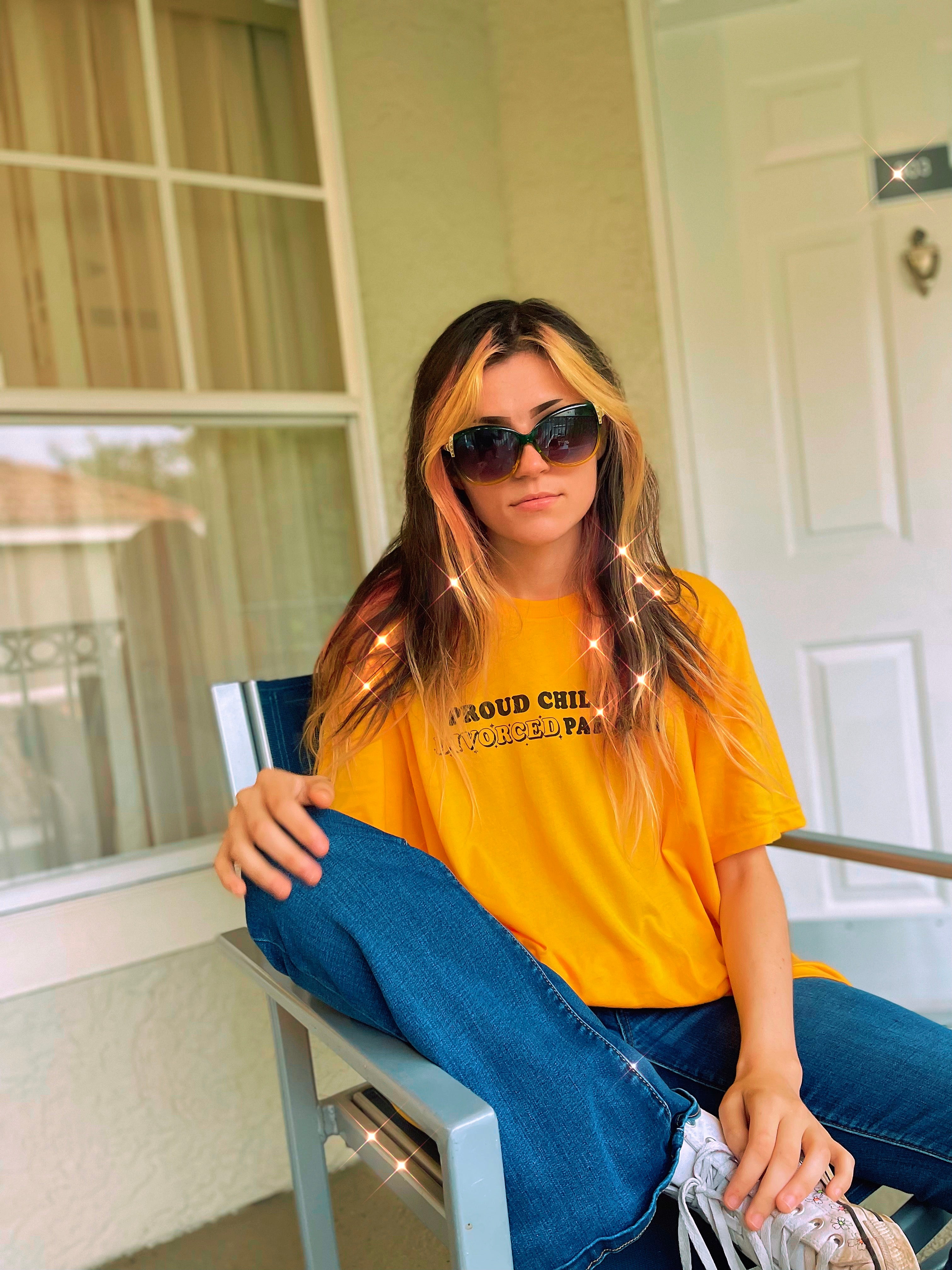 Woman wearing a bright yellow “Proud Child of Divorced Parents” graphic tee while sitting outdoors in sunglasses.