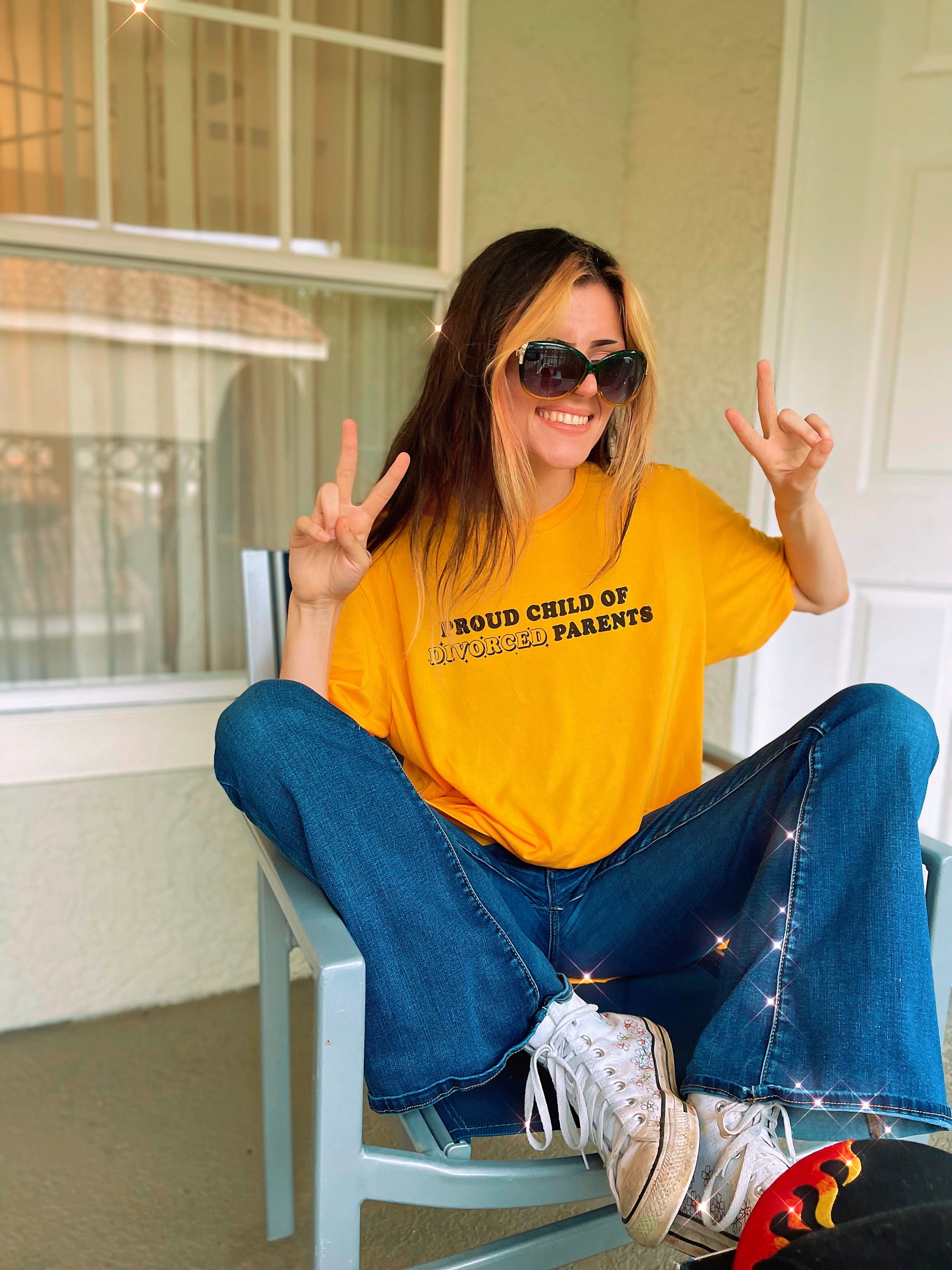 Woman smiling and throwing peace signs while wearing a bright yellow “Proud Child of Divorced Parents” graphic tee, jeans, and sunglasses.
