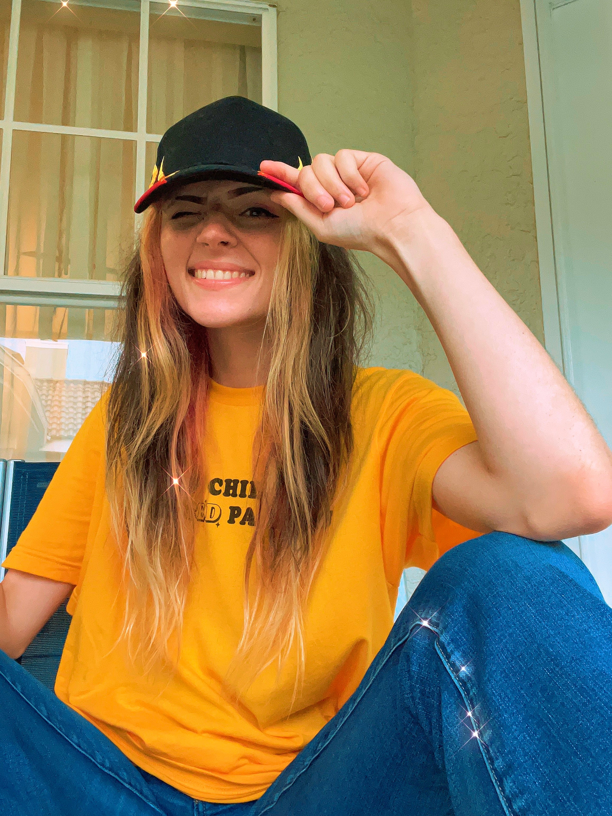 Woman smiling and tipping a hat while wearing a bright yellow “Proud Child of Divorced Parents” graphic tee and jeans.