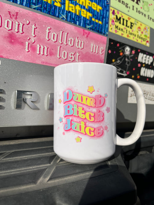 White ceramic mug with pink and blue text reading Dumb Bitch Juice surrounded by stars, placed outdoors on a black surface.