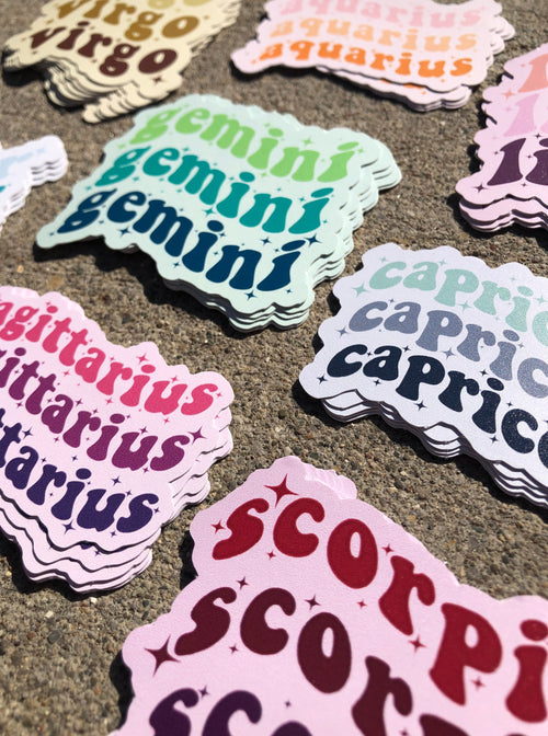 Stacks of colorful vinyl stickers for Gemini, Scorpio, Sagittarius, and Capricorn on a textured stone surface.