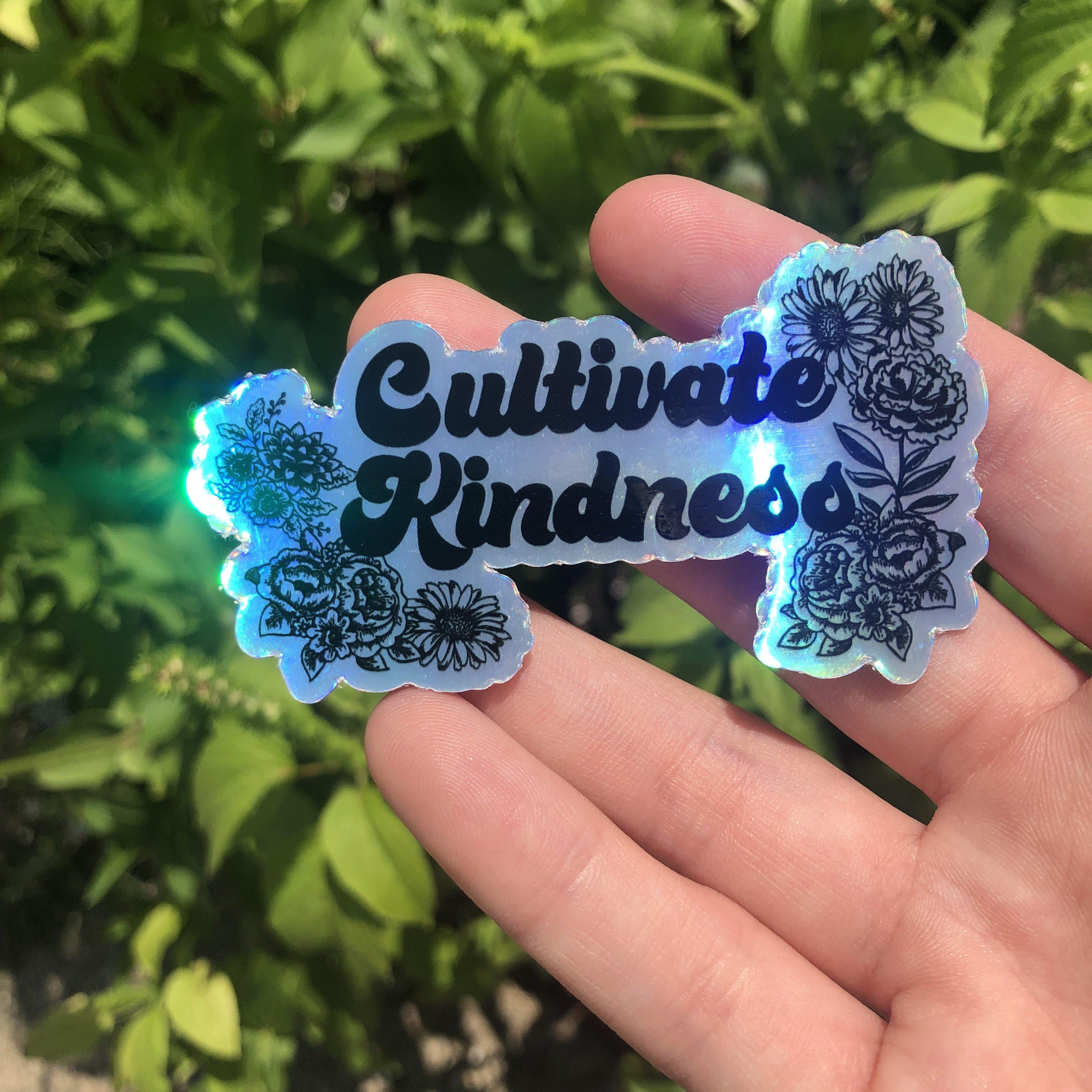 One holographic vinyl sticker held in a hand featuring black floral illustrations and the text Cultivate Kindness.