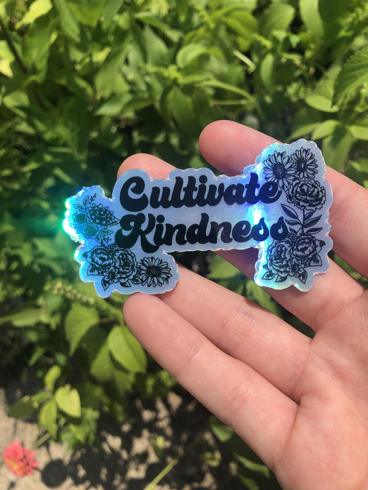 One holographic vinyl sticker held in a hand featuring black floral illustrations and the text Cultivate Kindness.