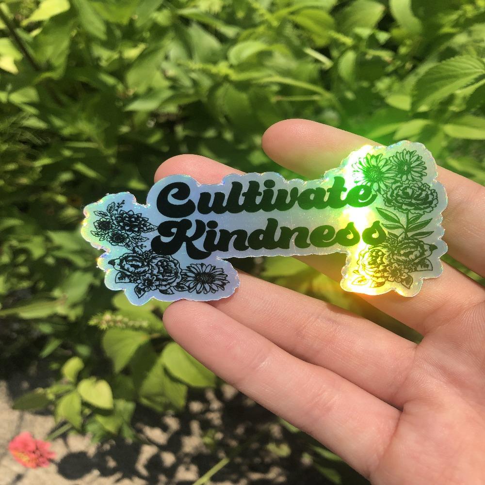 One holographic vinyl sticker with floral illustrations and the text Cultivate Kindness held in a hand.