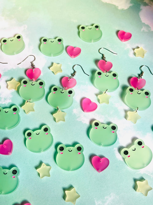 Green plastic frog face earrings with pink hearts and yellow stars scattered on a blue cloud background.