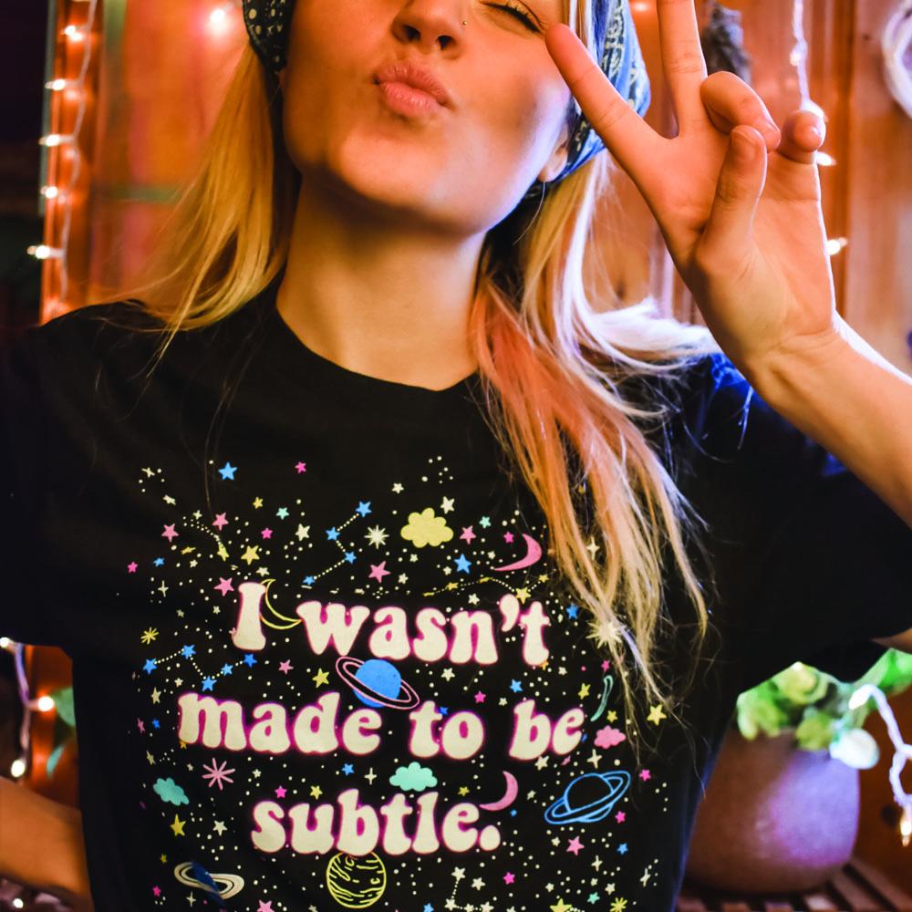 I Wasn’t Made to Be Subtle black graphic t-shirt with colorful stars, planets, and space-themed design, modeled by a person making a peace sign – bold retro cosmic statement tee – The Funnel Cake Tree.