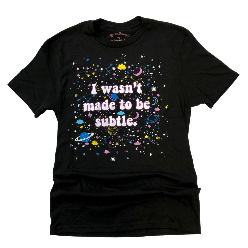 I Wasn’t Made to Be Subtle black graphic t-shirt with colorful stars, planets, and space design in pink, blue, and yellow – flat lay on white background – retro cosmic statement tee – The Funnel Cake Tree.